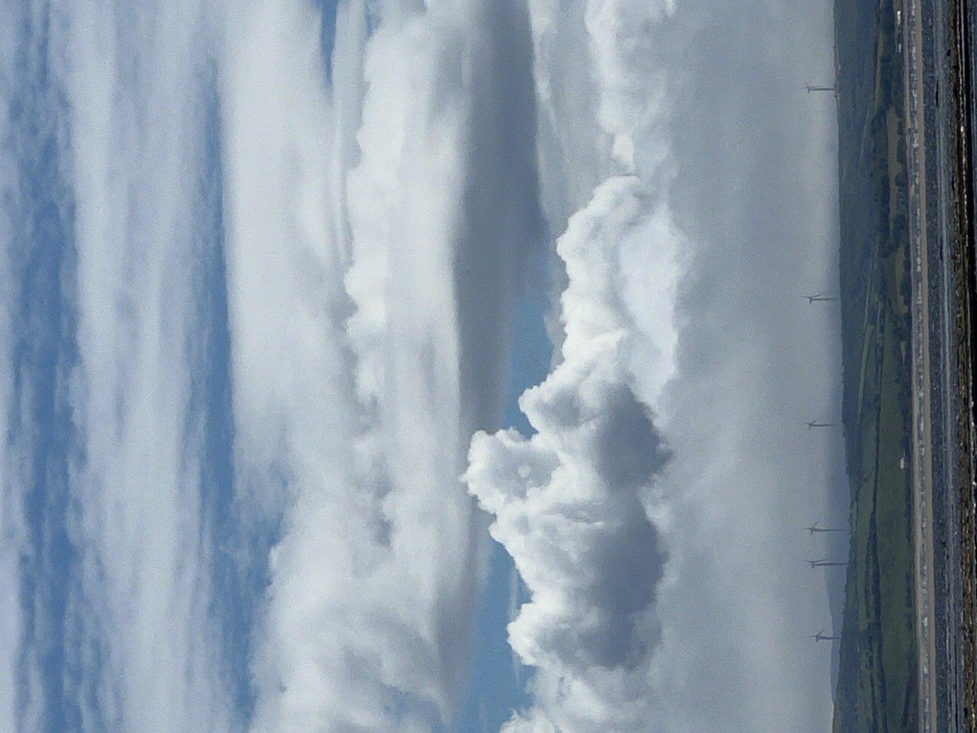 This media file, captured in Mountcharles, Ireland, presents a landscape dominated by a dramatic sky filled with large, billowy clouds. The clouds are primarily white and light gray, with some areas appearing darker, suggesting depth and variation in thickness. Patches of blue sky are visible between the clouds.
In the distance, several wind turbines are spaced out across a green, hilly landscape. The turbines are silhouetted against the sky and appear as tall, thin structures with arms. The ground below them is a patchwork of green fields, indicative of rural or agricultural land. A road or pathway runs along the right side of the image, bordered by what appears to be a body of water or a coastline.
The scene is captured vertically, with the horizon line running from left to right across the lower portion of the image. The lighting suggests it is daytime, likely late afternoon or early evening due to the diffused light and the potential for longer shadows, although these are not clearly discernible. The weather appears partly cloudy, with significant cloud cover.
There are no people or animals visible in the image. The overall mood is serene and natural, highlighting the power of the elements and the presence of renewable energy infrastructure within the landscape. No text is visible in the image.