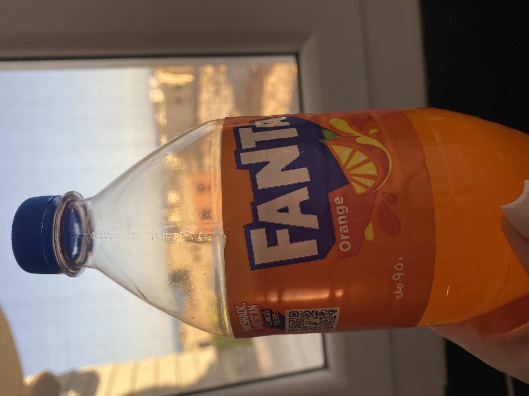 An orange Fanta bottle is held in a person's hand in front of a window. The bottle has a blue cap and a clear plastic body. The label on the bottle is predominantly orange with the Fanta logo in large white letters with a blue outline. The word "Orange" is written below the logo in smaller white text. To the left of the logo, there is a QR code and text that reads "SIP, SNACK, AND SCAN TO UNLOCK THE FUN". Arabic text indicating "90 ml" is visible on the lower part of the label. The background through the window shows a blurred view of buildings and a hazy sky, suggestive of an outdoor setting in a city. The lighting indicates it is daytime, possibly late afternoon given the warm tones. The environment appears to be indoors, with the window framing the outdoor scene. The image is taken from a low angle, focusing on the Fanta bottle. This scene could be located in Sharm El Sheikh, Egypt, given the presence of Arabic text and the commonality of such bottled beverages in tourist and residential areas.