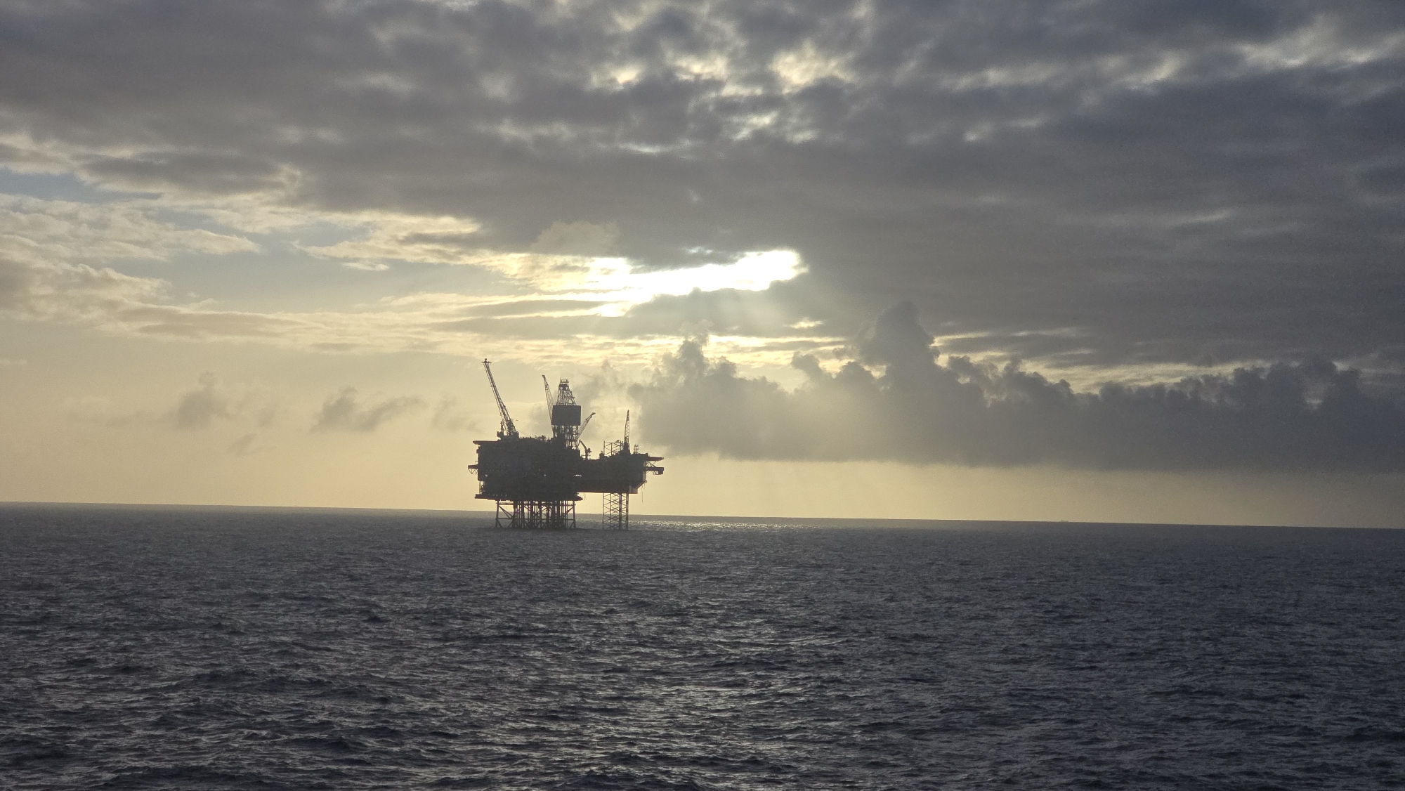 The image shows an oil rig platform in the North Sea, likely near Randaberg, Norway, given the typical location of these platforms. The platform is silhouetted against the background of the sky and sea, with the sun partially obscured by clouds, creating a dramatic lighting effect with rays of light breaking through. The sea is choppy. There are no people visible in the image.