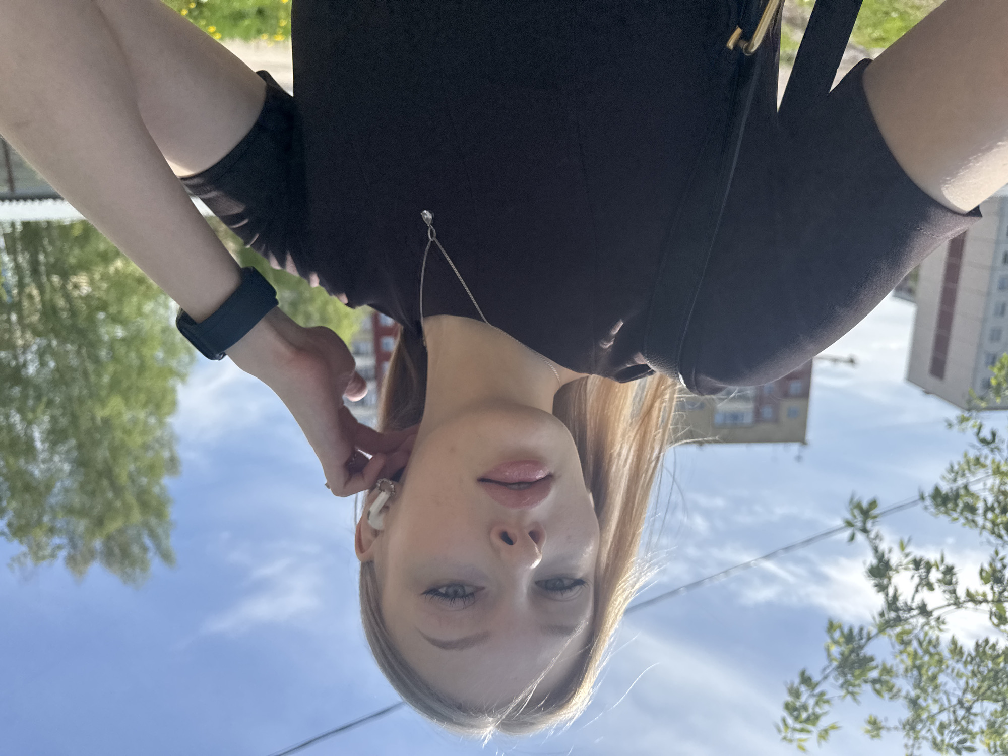 A medium close-up perspective captures a fair-skinned woman with blonde hair from a low angle, appearing to hold the camera above her head. The background reveals a clear blue sky with scattered white clouds, and glimpses of urban elements. To her left is a tree with lush green foliage; to her right, an apartment building typical of urban areas in Russia. She is wearing a dark-colored top and a black fitness tracker on her left wrist. An earbud is visible in her right ear. A silver necklace hangs from her neck. The location is identified as the city of Perm, Russia based on architectural styles of buildings and the type of trees in the image.