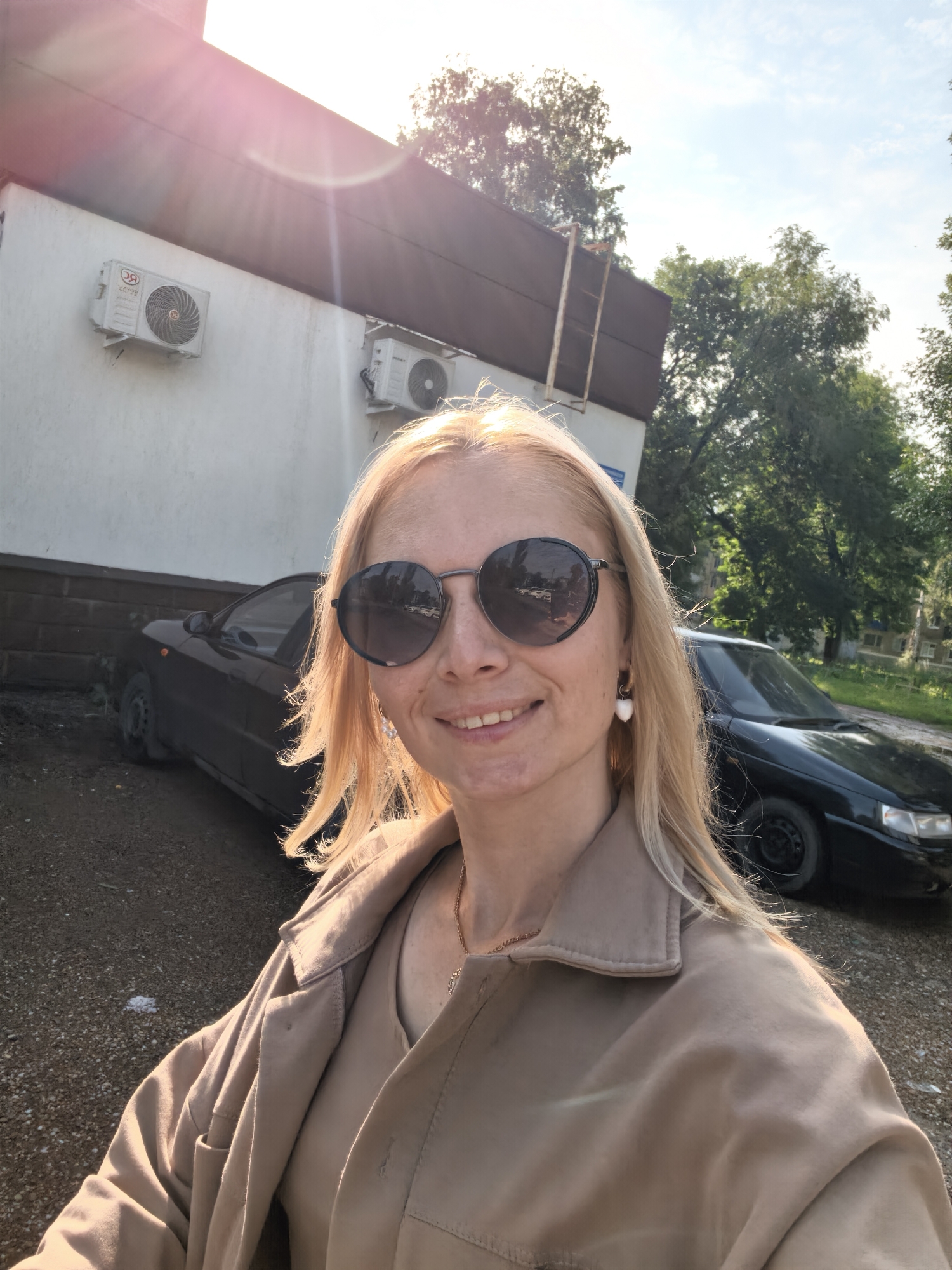 This is a selfie taken outdoors in Sterlitamak, Russia, featuring a fair-skinned woman with blonde hair and sunglasses. She is wearing a tan jacket and a necklace. In the background, two black cars are parked. One is a dark sedan behind the woman, and another is partially visible further back. A building with two air conditioning units mounted on its white exterior wall is also present. Trees and a bright sky suggest a daytime setting, possibly with the sun setting or rising due to the lens flare and warm lighting. The ground is a mix of gravel and dirt.