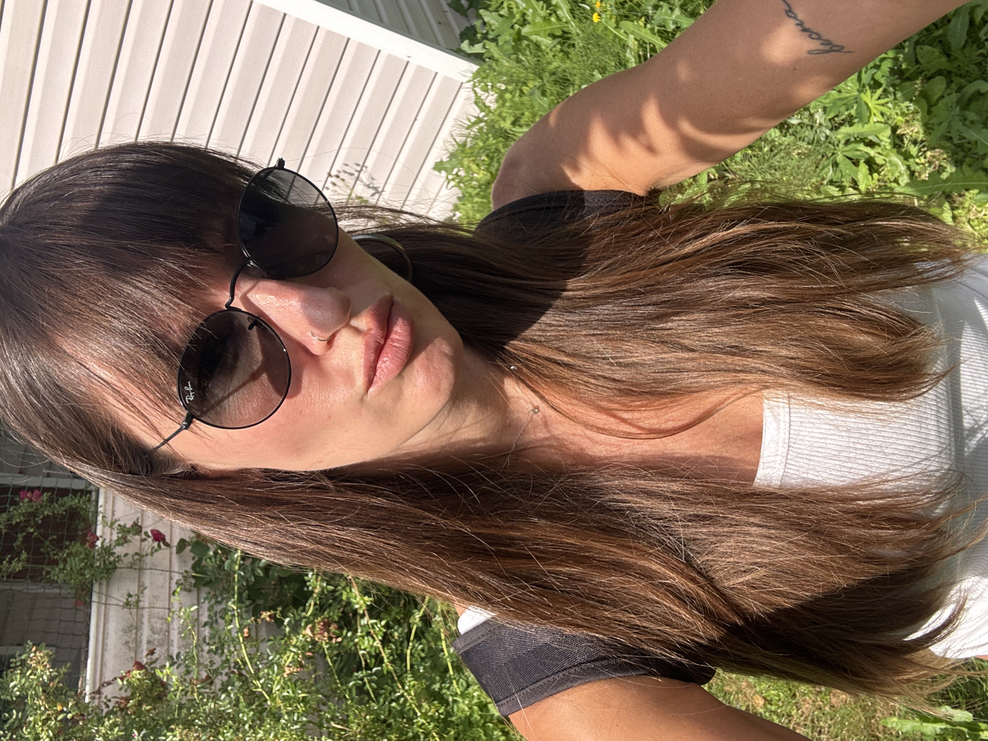 In the city of Krasnaya Presnya SNT, Russia, a woman with long brown hair and bangs is taking a selfie outdoors. She is wearing a white tank top, sunglasses, and a nose ring. A tattoo is visible on her right arm. She is standing in front of a light-colored building with horizontal siding, and lush green vegetation surrounds her. The lighting suggests it is a sunny day.