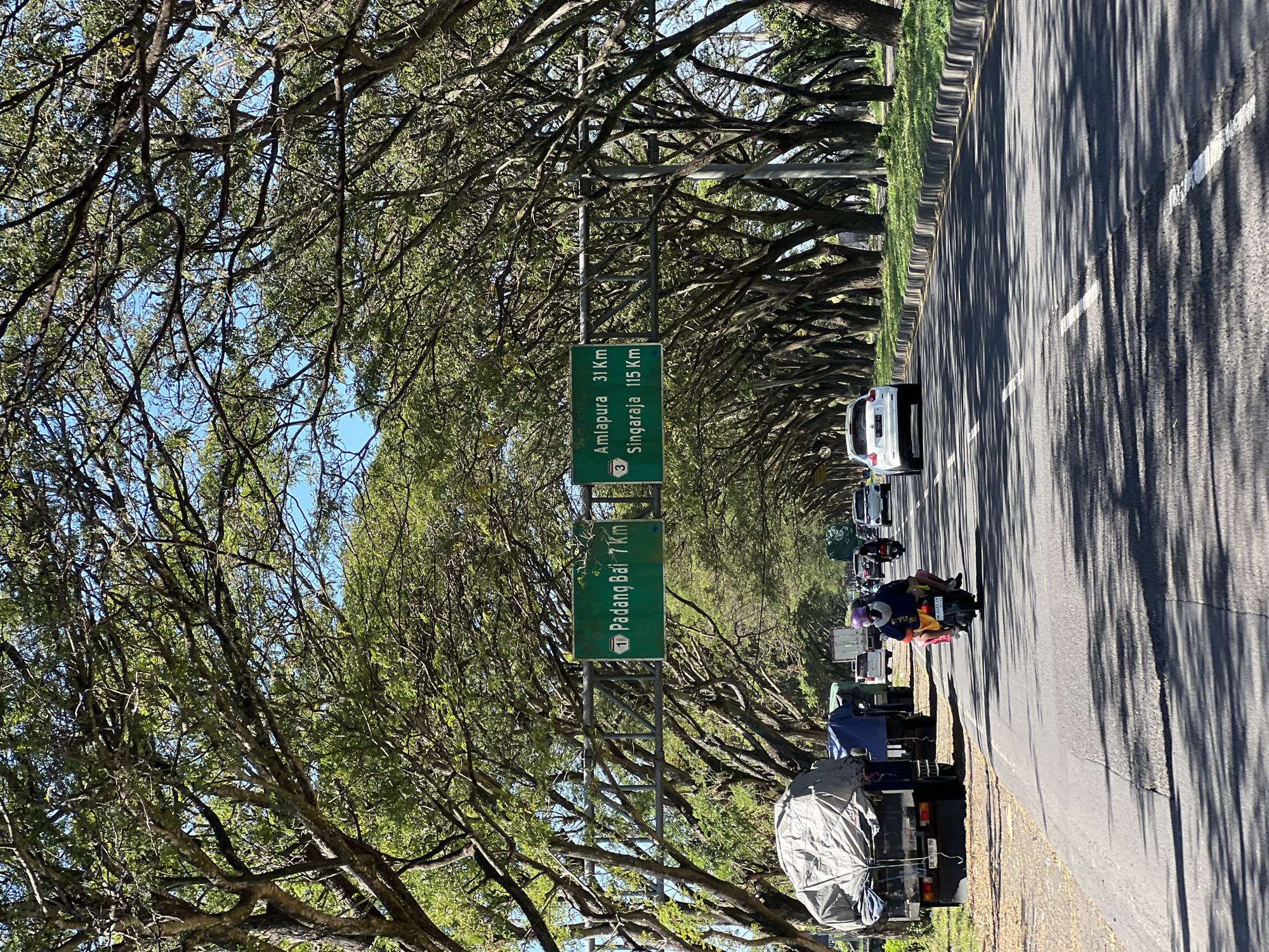 In Dawan, Indonesia, this image captures a sun-drenched, tree-lined road. A two-lane asphalt road stretches into the distance, bordered by lush green grass and densely planted trees whose branches interlace overhead, forming a natural tunnel. Two green road signs are suspended from a metal structure above the road. The left sign indicates "1 Padang Bai 7 Km" and the right sign reads "Amlapura 31 Km" and "Singaraja 115 Km."
Traffic moves in both directions. On the left shoulder, a large truck covered with a gray tarp is parked, alongside other parked vehicles. In the foreground, a motorcycle with two occupants is traveling on the right lane. The driver wears a blue shirt and a pink helmet, while the passenger is dressed in a yellow and pink outfit. Several cars are visible further down the road, with a white sedan in the right lane and other vehicles in the left lane. The sky is a clear, bright blue, with dappled sunlight casting shadows from the trees onto the road surface. The overall scene depicts a typical day on a scenic highway in a tropical region.