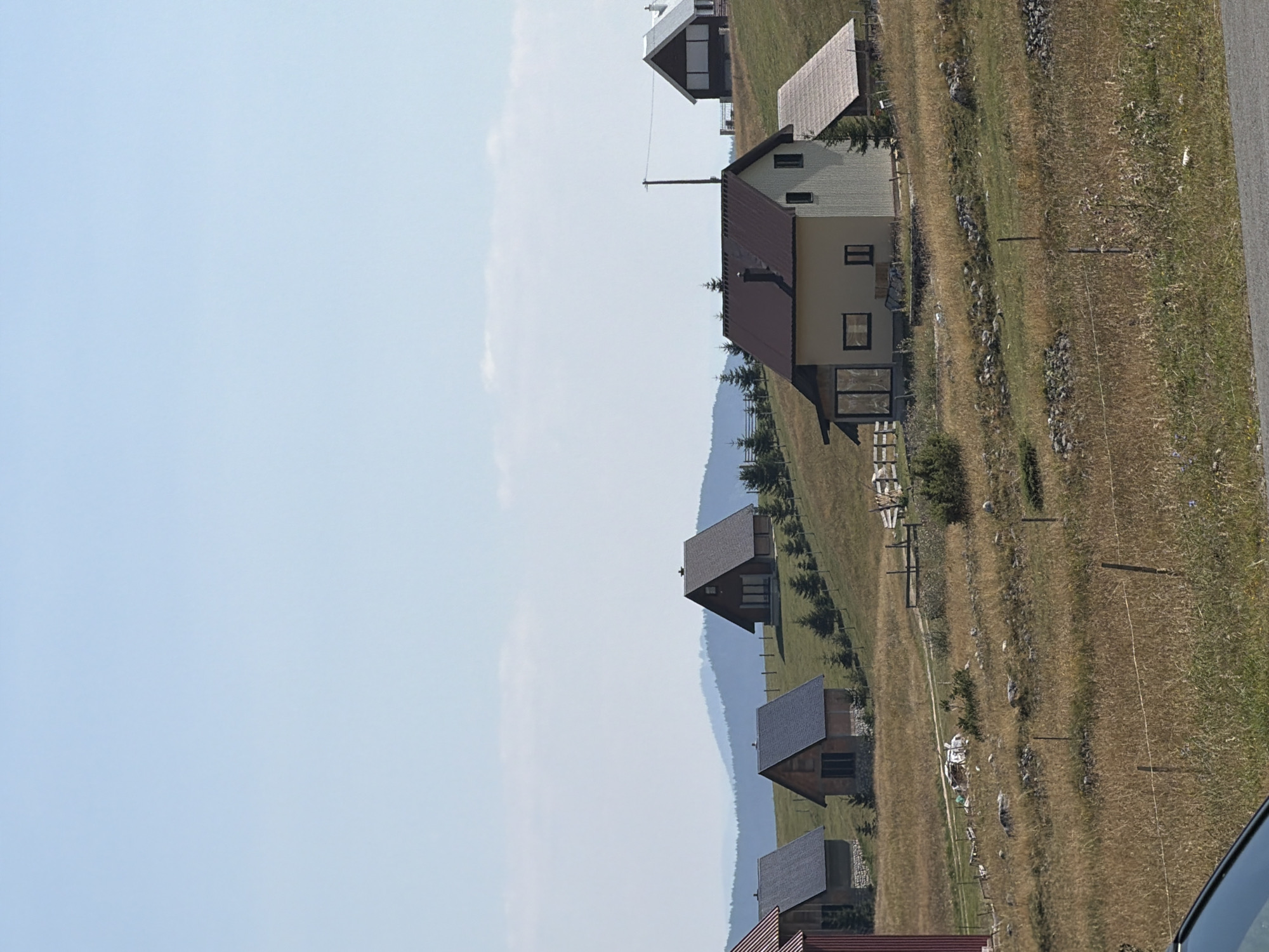 The image depicts a rural landscape in Virak, Montenegro, featuring several houses scattered across rolling hills. The foreground shows a portion of a dark-colored vehicle, indicating a road or driveway. The middle ground is dominated by dry, golden grass and scattered stones, with a fence running across the scene. A variety of small to medium-sized houses with pitched roofs are visible on the hillside, some with visible windows and chimneys. Some of these houses appear to be made of wood and stone, while others have lighter-colored siding. A few small evergreen trees are planted around the houses and along the fence line. In the background, more distant hills are visible, fading into a hazy blue due to atmospheric perspective. The sky is a clear, pale blue. The overall impression is of a tranquil, undeveloped rural setting.