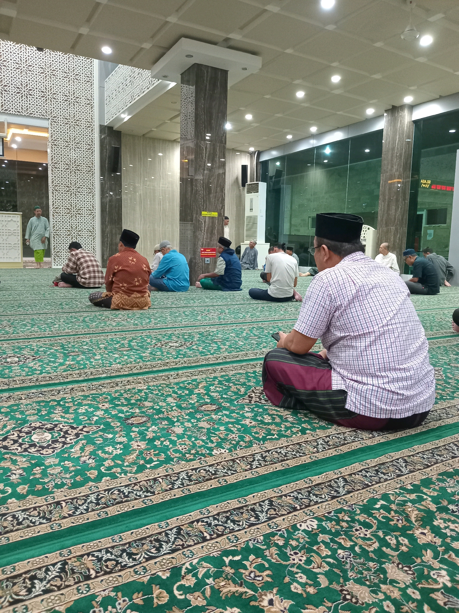 The image depicts an interior view of a mosque in Randuagung, Indonesia, likely during prayer time. Several men are seated on a green carpet with intricate patterns, facing away from the camera. They are dressed in traditional Islamic attire, including peci caps, sarongs, and long-sleeved shirts. In the foreground, a man wearing a checkered shirt and a maroon sarong is seated with a mobile phone in his hand. The mosque's architecture features patterned wall panels and large windows, with ceiling lights illuminating the space. An air conditioning unit is visible on the left side of the frame. The overall atmosphere suggests a gathering for religious observance.