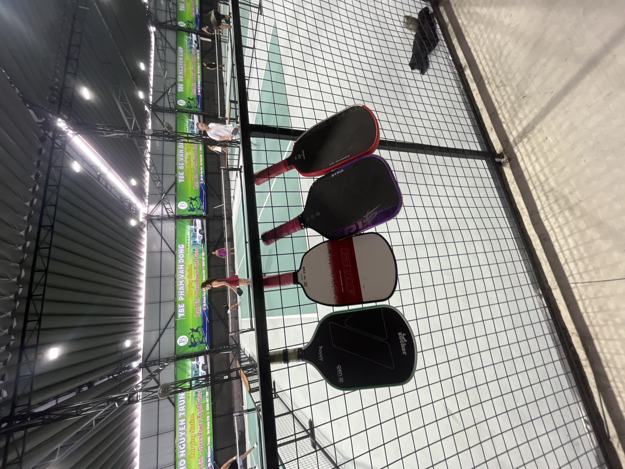This image depicts an indoor pickleball facility in Đà Nẵng City, Vietnam, specifically located near 291-311 Ho Nguyen Trung as indicated on the background banners. The perspective is from a spectator area, looking across multiple pickleball courts through a black wire mesh fence.
In the immediate foreground, four pickleball paddles are suspended from the top rail of the wire fence. From left to right, these include a black paddle with green trim ("Zocker," "gen 2," "happy" visible upside down), a white paddle with a red abstract design ("GRAF" visible), a black paddle with purple accents ("SYPIK" visible), and a black paddle with red accents, also "SYPIK" branded. The paddles feature pink and red handle wraps. The floor beneath the fence is light gray concrete, with shadows cast across its surface.
Beyond the fence, several vibrant light green pickleball courts are visible, each demarcated by white lines and separated by white nets and low dark perimeter fencing. Multiple individuals are engaged in play on these courts. On the court to the right of the frame's center, a woman in a red dress holds a paddle, facing left. Further back on the same court, another woman in a purple outfit stands. On the adjacent court to the left, a man in a white t-shirt and dark shorts holds a paddle, facing right, also actively playing. Additional players are visible on courts further in the background. Wooden benches are positioned along the court perimeters.
Dominating the background are multiple large green banners affixed to the back wall. These banners advertise a "Charging Station" with the slogan "Thanh Bình - Green Power, Bright Future" and feature the "VINFAST" logo. Crucially, they list street names such as "TBE PHAM VAN DONG," "TBE VO NGUYEN GIAP," "TBE LE VAN DUYET," and "TBE HO NGUYEN TRUNG," confirming the specific location within Đà Nẵng City, Vietnam, along with the address "291-311 Ho Nguyen Trung." The banners incorporate graphics of people playing a sport and white electric vehicles.
The facility's ceiling consists of a dark gray, corrugated metal roof, supported by an exposed framework of black metal trusses and beams. Numerous bright white LED strip lights are mounted along these structures, providing uniform illumination for the courts. The overall environment suggests a well-maintained, dedicated indoor sports venue. In the lower right corner, behind the fence, a black bag rests on the concrete floor.
