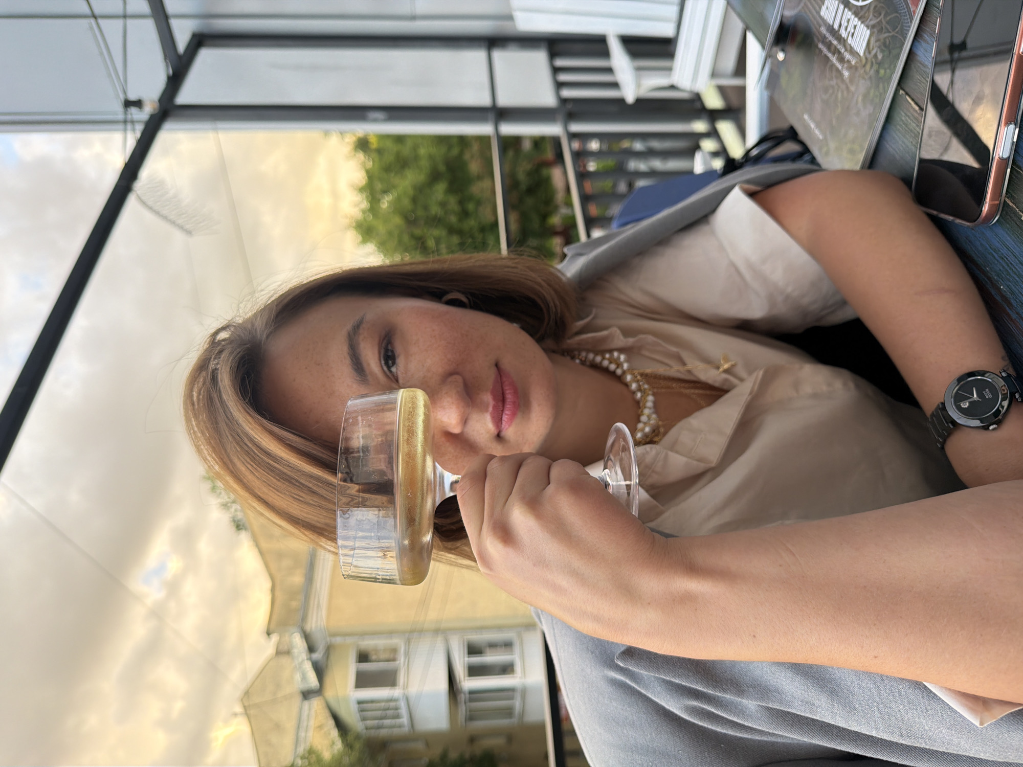 A medium shot captures a woman holding a glass of champagne up to her right eye, obscuring it from view. She has short, auburn hair and is wearing a light-colored blouse layered under a gray jacket. A pearl necklace adorns her neck, and a dark wristwatch is visible on her left wrist. The background shows a building with yellow facades and balconies, suggestive of an urban environment. The woman is seated outdoors, with a railing and chairs visible to her right, indicating she is likely at a restaurant or cafe with an outdoor seating area. The sky above is partly cloudy with a warm, golden hue, suggesting it is either sunrise or sunset. The scene appears to be in the city of Izhevsk, Russia.