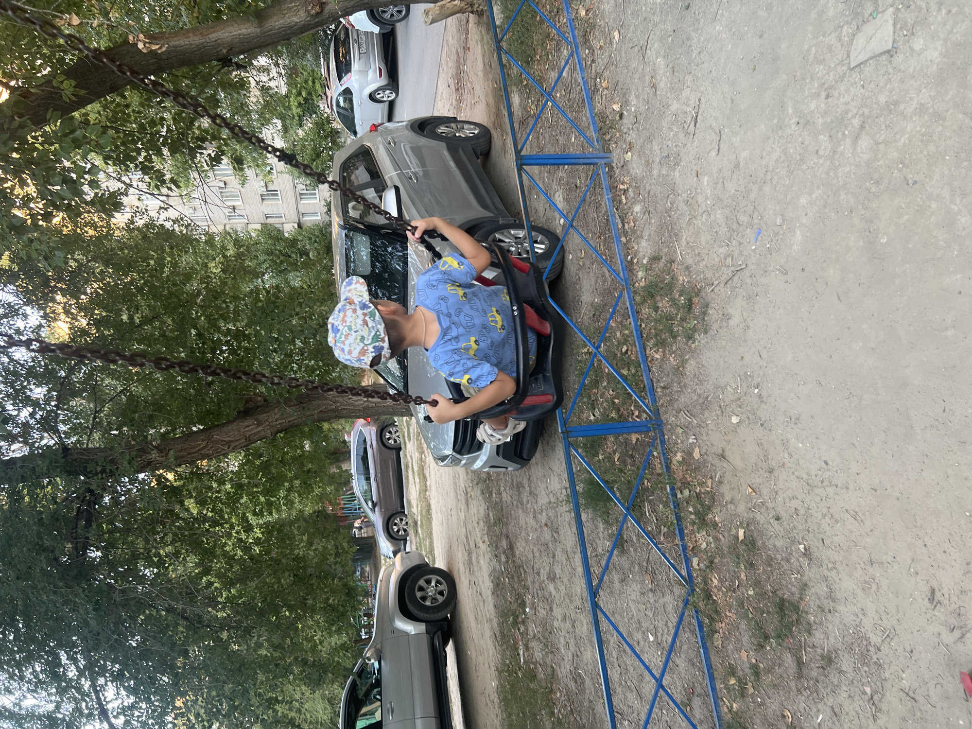 A young boy, wearing a blue t-shirt with yellow car motifs and a cap with a dinosaur pattern, sits on a swing. He is captured from behind, holding the swing chains and looking towards a silver SUV parked in front of him. The swing set is in a dirt and gravel area, bordered by a blue metal fence. In the background, several parked cars, trees, and a multi-story apartment building are visible, suggesting an urban residential setting. The lighting indicates it is daytime, possibly late afternoon, with some sunlight filtering through the trees. The scene appears to be in a courtyard or playground area of a city.