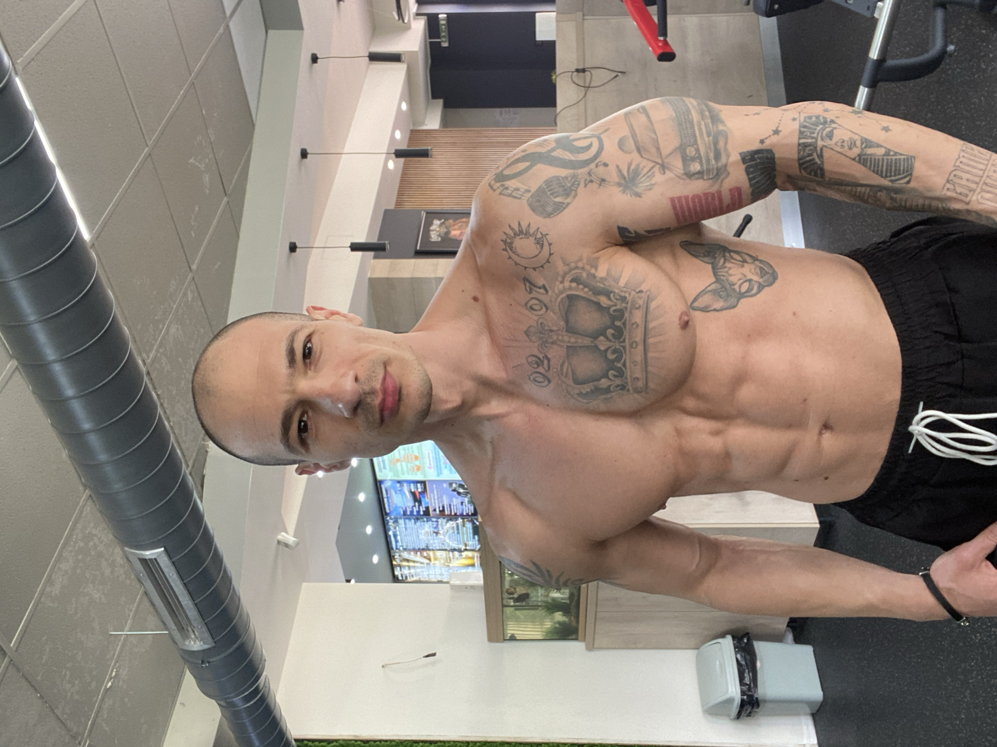 A shirtless man with visible tattoos stands in what appears to be a gym or health club. He faces the camera directly. He is bald. The environment includes exercise equipment visible in the background and part of the ceiling and walls, suggesting an indoor setting. A restaurant sign with a menu is visible. The location cannot be confirmed as the city of Pleven, Bulgaria, based on the image alone. No specific actions or interactions are apparent other than the man posing for the image.