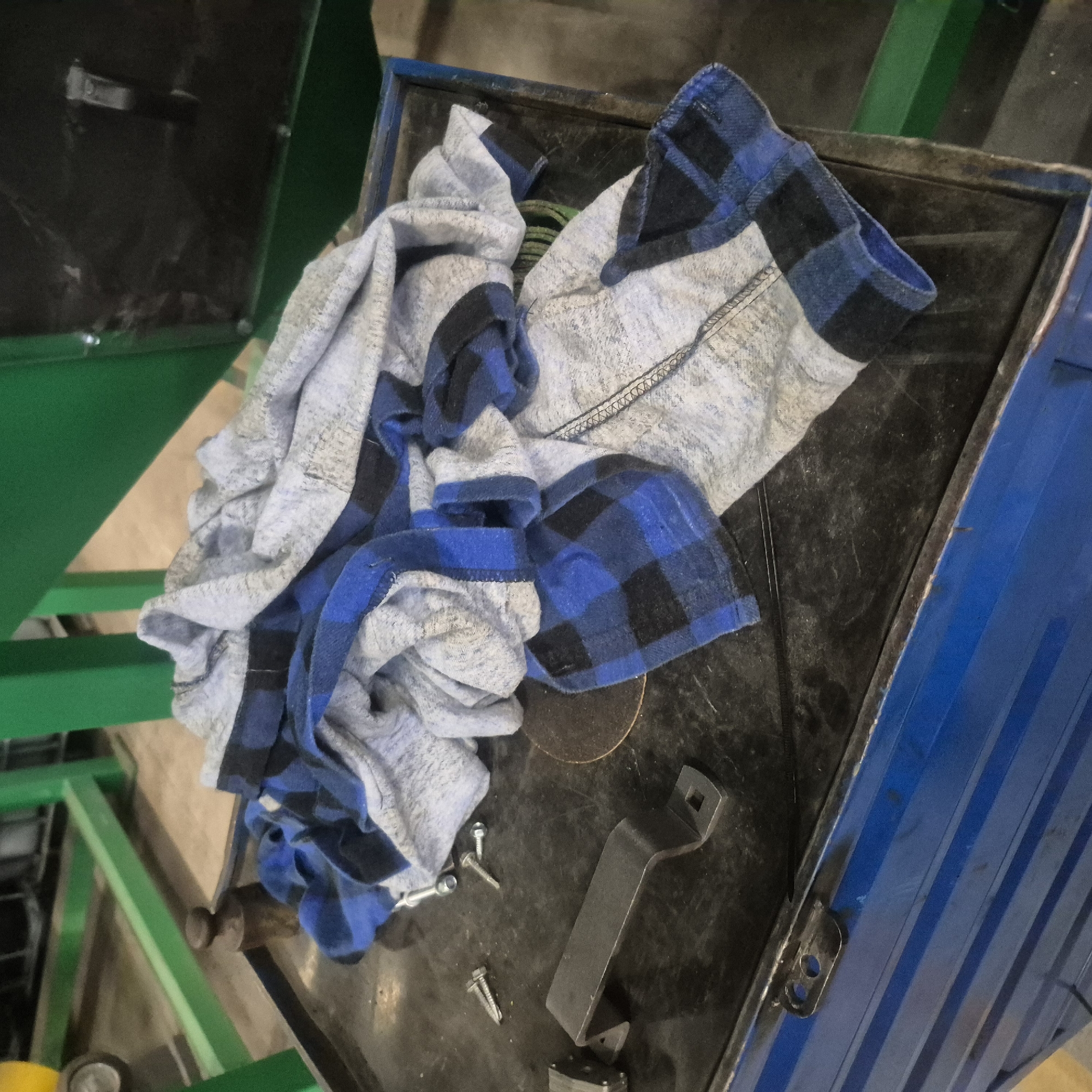 A pile of crumpled clothing, featuring a blue and black plaid pattern mixed with grey fabric, rests on a black surface. Adjacent to the clothing are several screws and a metal bracket. In the background, industrial green metal structures are visible. The scene is set within a workshop or industrial environment. The image is taken from a high angle, looking down at the objects on the black surface.