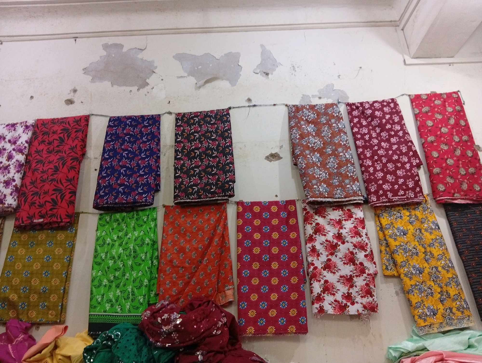 The image shows a row of fabrics hanging on a line against a distressed, off-white wall. The fabrics are of various colors and patterns, including floral prints and geometric designs. Some are folded, while others are displayed full length. In the foreground, several piles of colorful fabrics are visible, with some appearing to be silk or satin. The setting appears to be a market or shop in Larkana, Pakistan, likely a textile stall. The wall exhibits peeling paint and some imperfections, suggesting an aged structure. There are no people visible in the image.