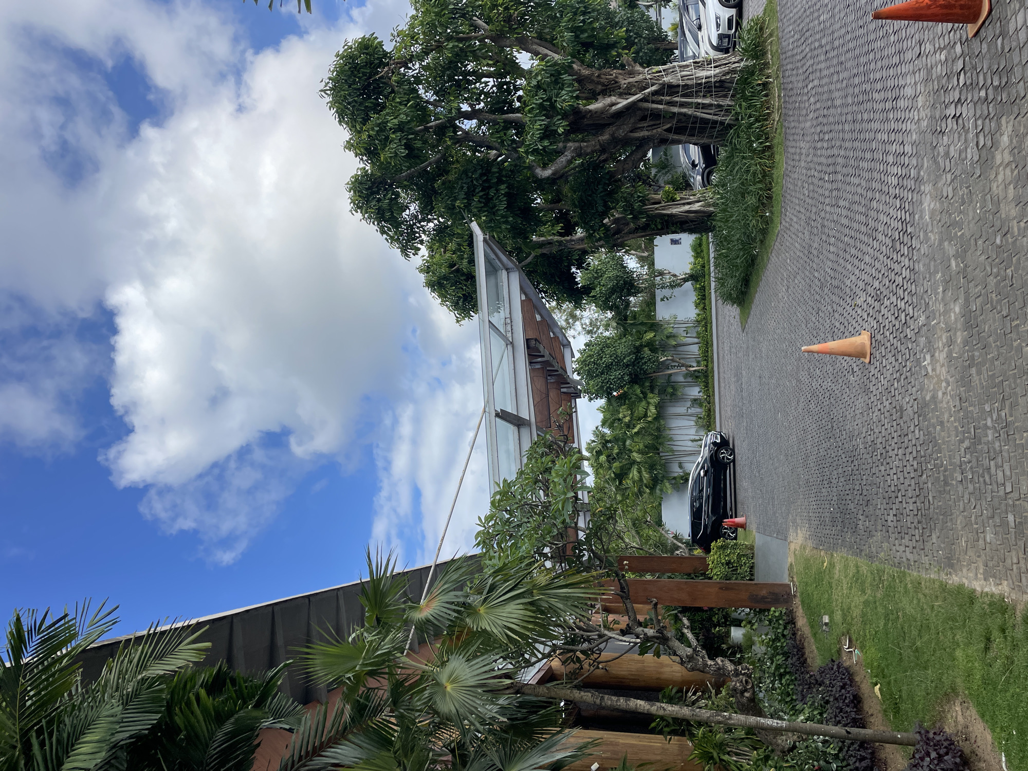 The image presents an outdoor scene, likely a paved driveway or walkway with interlocking bricks, bordered by greenery. Two orange traffic cones are placed along the edge of the pathway. A large, mature tree with aerial roots is visible, casting shadows on the brick surface. Several parked cars are near the tree, including a dark-colored car beside the driveway and white cars partially visible. Buildings are also present, with glass walls and wooden structures that are lined with foliage. The sky is partly cloudy, with patches of blue. The perspective is angled upwards, with the camera positioned relatively low. The location is presumed to be in Unknown, Unknown.
