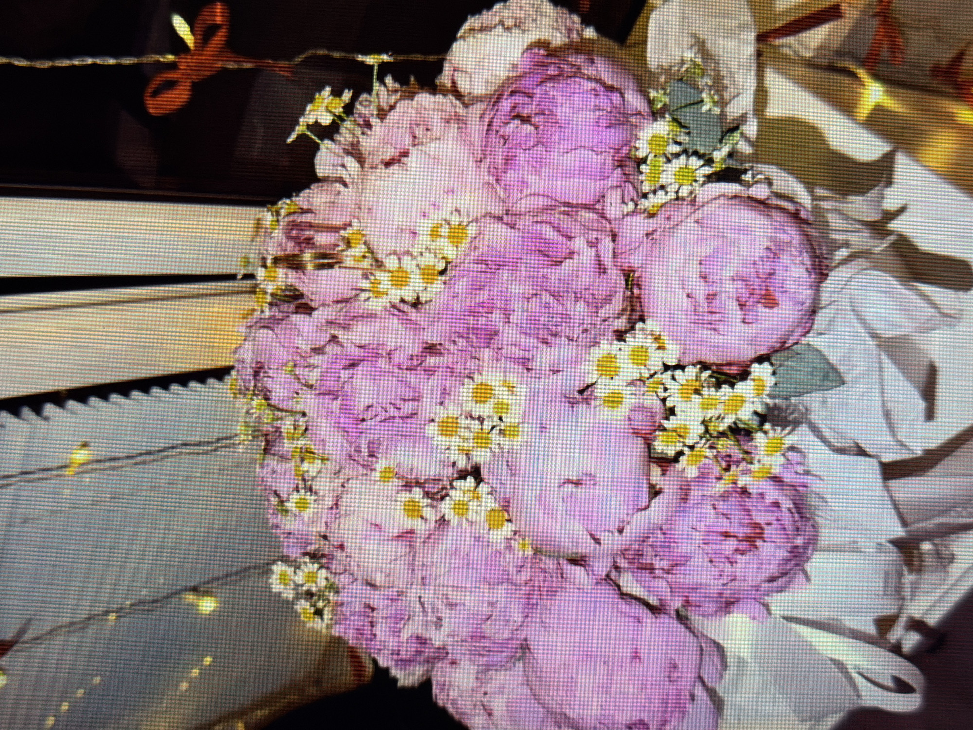 A floral arrangement of pink peonies and white daisies is presented on a white cloth. The background consists of a light-colored, textured surface, possibly wood, with strands of small, yellow lights draped above the flowers. To the right, a dark background with a red ribbon tied in a bow is visible. The scene is illuminated, casting shadows from the flowers and ribbon onto the surrounding surfaces. There are no people or animals present in the image. The image does not contain information about its location in the city of Sirius, Russia.