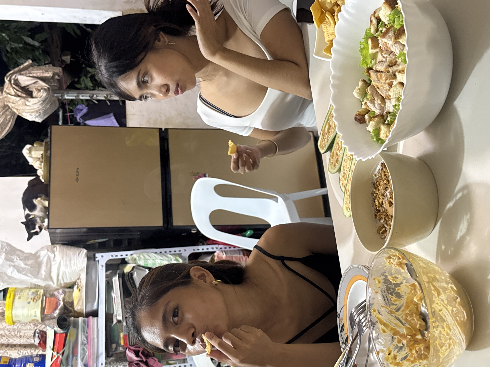 Two young women are seated at a table with food. The woman on the left is wearing a black spaghetti strap top and is eating a cracker. The woman on the right is wearing a white t-shirt with a black strap visible and is holding a cracker, looking to the side. On the table, there is a bowl of salad with chicken and croutons, a bowl of granola or cereal, sliced zucchini, and a bowl with remaining food. In the background, there is a refrigerator and shelves with various items. A cat is perched on top of the refrigerator. The scene appears to be indoors, possibly a dining area, and the presence of local food items suggests it is in Davao City, Philippines.