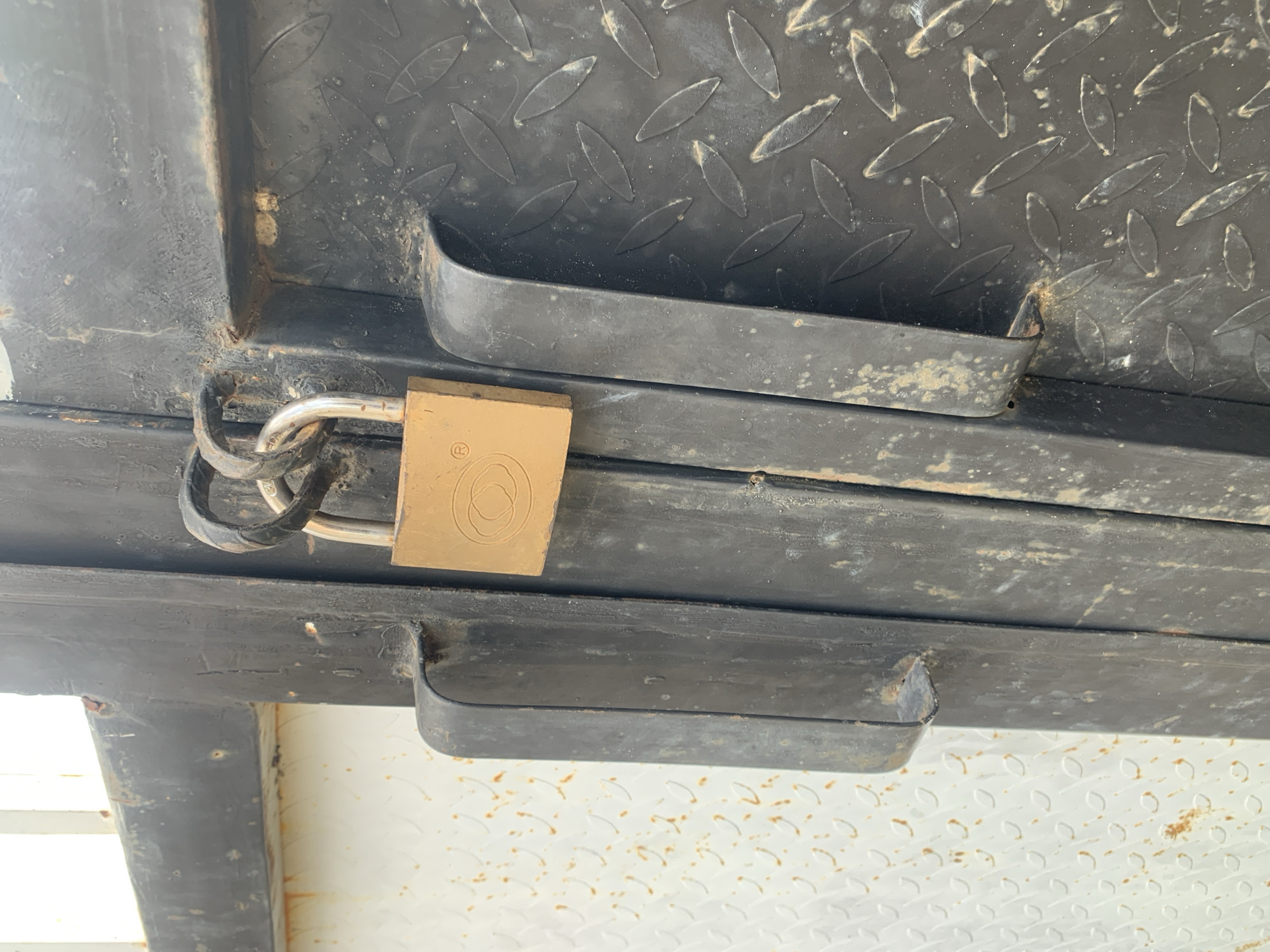 A close-up, eye-level shot shows a weathered brass padlock hanging from two interlocking metal loops on a dark, industrial-style metal door. The padlock is rectangular with a circular logo on its face. The door's surface is textured with a diamond plate pattern and shows signs of rust and dirt. A portion of a white, textured surface with orange rust stains is visible to the left of the door. Two vertical black handles are attached to the door, one on either side of the padlock. The overall environment appears to be outdoors in an urban or industrial setting, suggesting a secure entry point that is currently locked.
