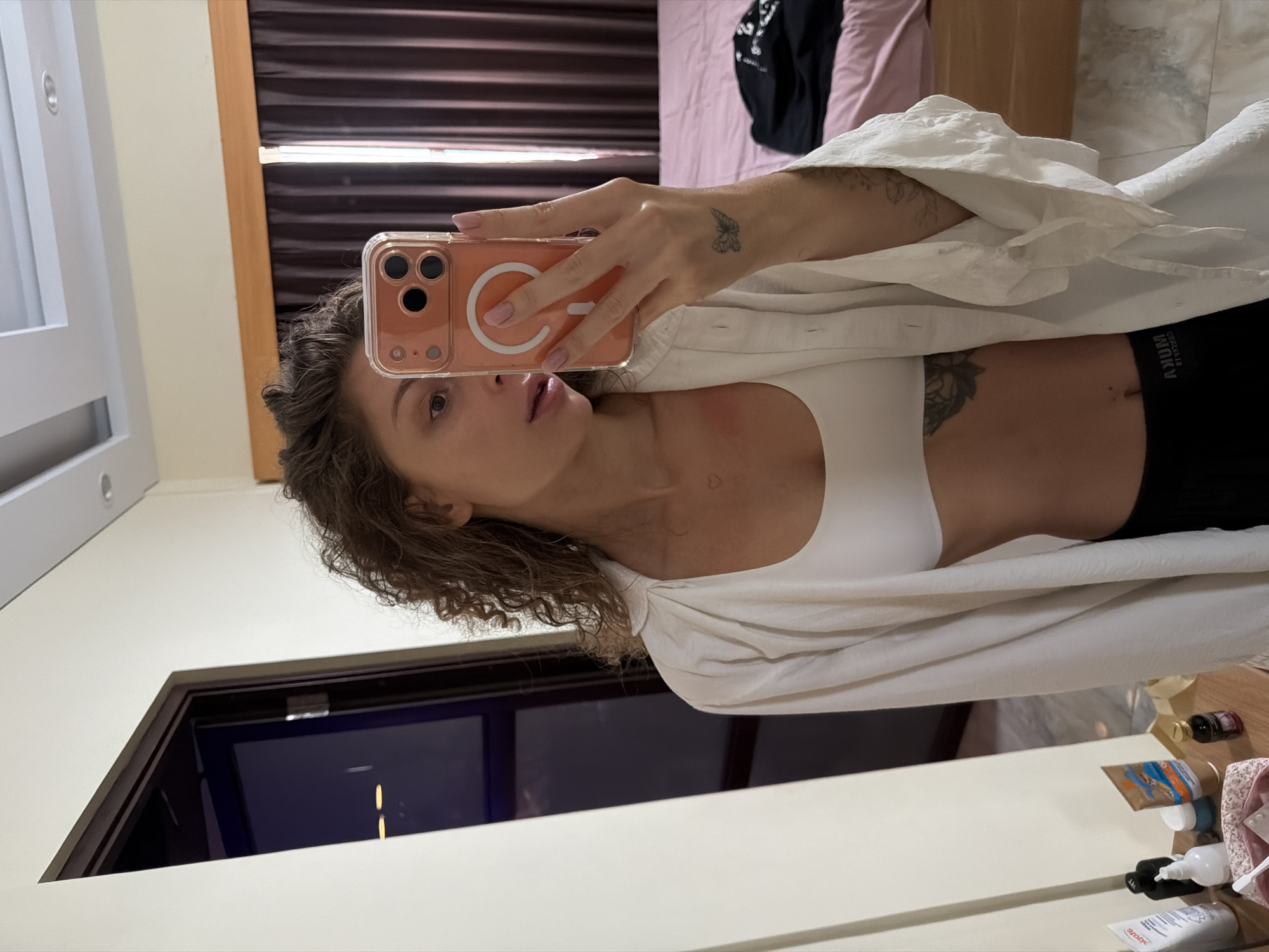The image, originally rotated 90 degrees counter-clockwise, depicts a woman taking a mirror selfie in an indoor setting, likely a bedroom, in North Kuta, Indonesia.
The primary subject is the woman, who appears to be standing or sitting upright, holding a smartphone with an orange, clear case and a visible MagSafe ring in her right hand to capture her reflection. She has curly brown hair, and her expression is neutral, with her lips slightly parted as she looks towards the phone. She is wearing a white sports bra or crop top beneath an unbuttoned, oversized white shirt with textured fabric. Black shorts or underwear with a white waistband are visible below her waist.
Notable details include several tattoos: a small butterfly on her right hand/wrist, a tiny heart on her neck, and a larger, possibly floral or mandala-style tattoo on her left rib cage/torso area. Her fingernails are painted a light pink or nude color.
The background, reflected in the mirror, shows parts of a room. To the left, there's a white cabinet or furniture piece. In the upper background, dark brown blinds or curtains are visible, with a bed featuring pink bedding and a black item of clothing casually placed on it. To the right, a dark wooden doorframe reflects a dark interior, possibly a hallway or another room. Along the bottom edge of the mirror, various toiletries and bottles are reflected on a surface, including one with "AROMATIKA" visible on its label.
The visible text "CRITICAL CROP BY AKUNKI" is clearly seen on the white waistband of her black shorts/underwear. The overall atmosphere is casual and personal, consistent with a moment captured in a private living space. No specific time of day or weather cues are evident.
