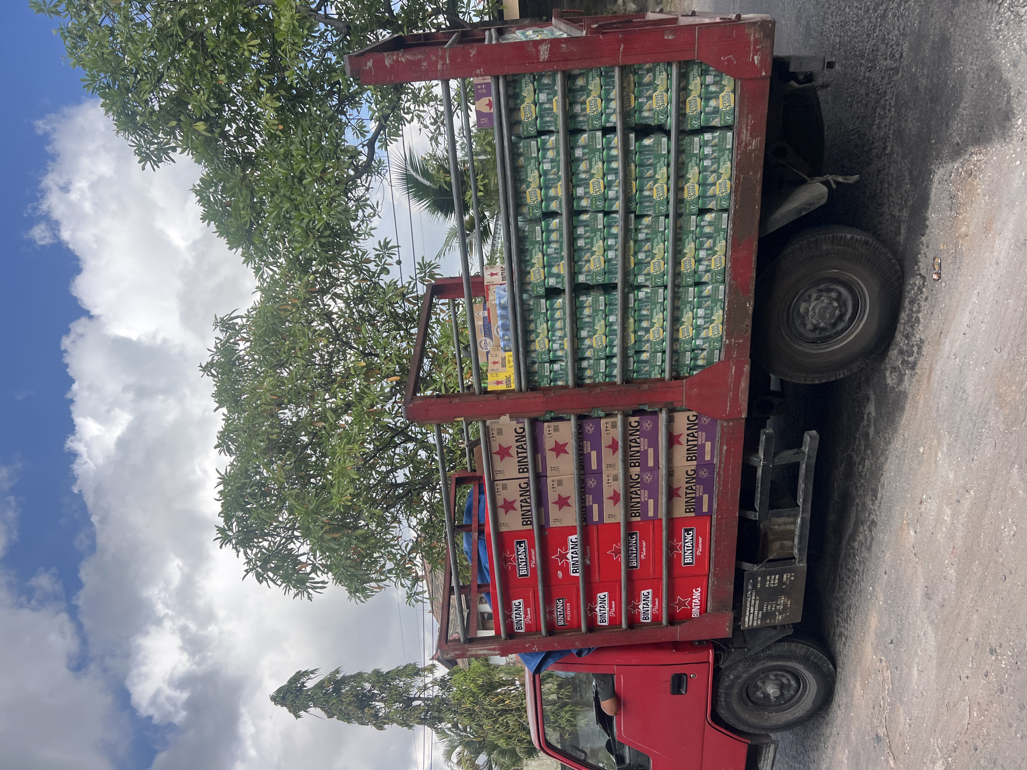 A red truck is parked on a paved road in Denpasar, Indonesia, its bed filled with cases of beverages. The truck is loaded with multiple stacks of boxes, predominantly displaying the "BINTANG" logo and some with "GREEN SANDS LIME APPLE" branding. The "BINTANG" boxes are primarily red and purple, while the "GREEN SANDS" boxes are green. A person's arm is visible through the driver's side window, indicating the driver is likely present. The background shows lush green trees, a tall, slender tree on the left, and a blue sky with scattered clouds. The scene appears to be captured during the daytime.