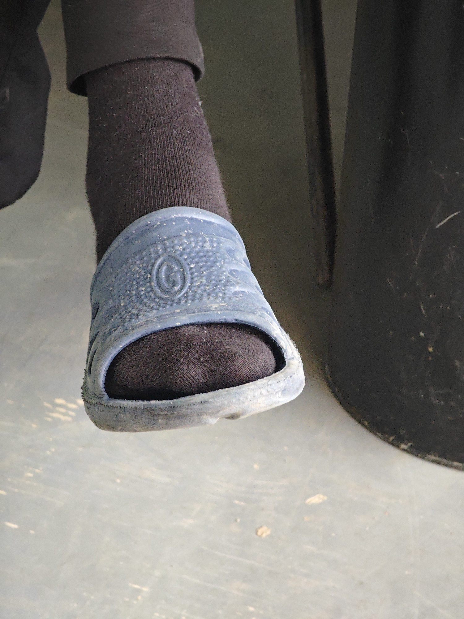 The image shows a close-up view of a person's left foot wearing a dark sock and a blue sandal. The sandal is made of plastic and has a textured surface with a circular emblem. The foot is resting on a light-colored, possibly concrete, surface. In the background, a dark, cylindrical object and a vertical dark object, possibly a chair or table leg, are visible. The scene appears to be indoors or in a sheltered outdoor area. There is no specific information within the image that indicates a location in Krasnoyarsk, Russia.