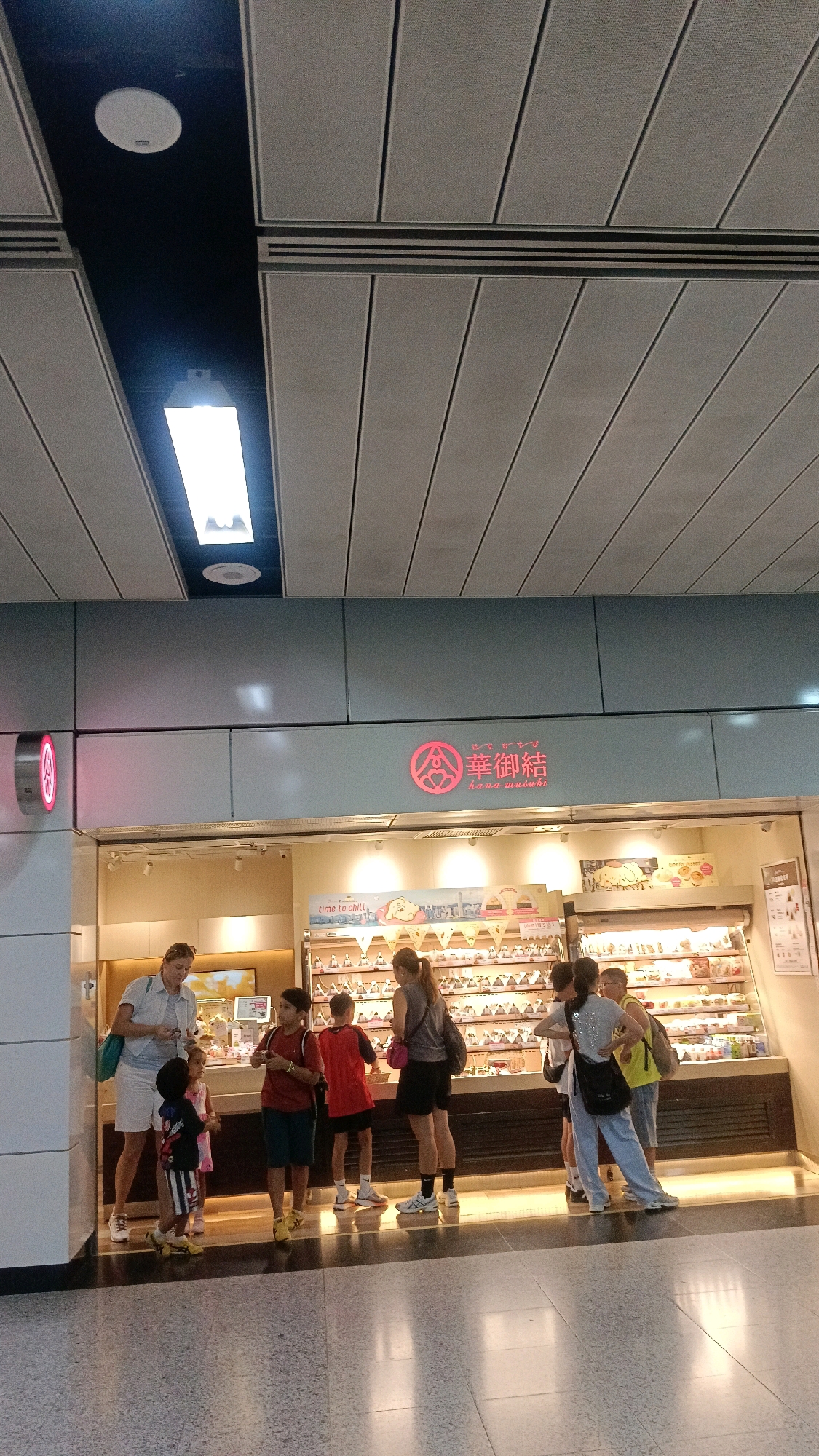 A group of people, including adults and children, stand in front of a food stall in a shopping area. The stall displays a variety of food items in a refrigerated glass case. A prominent sign above the stall features Chinese characters and the English words "HANA MUSUBI". The individuals appear to be browsing or waiting in line. The environment suggests an indoor public space, possibly a mall or a transportation hub, with tiled flooring and a modern ceiling. The scene is brightly lit by overhead lighting.