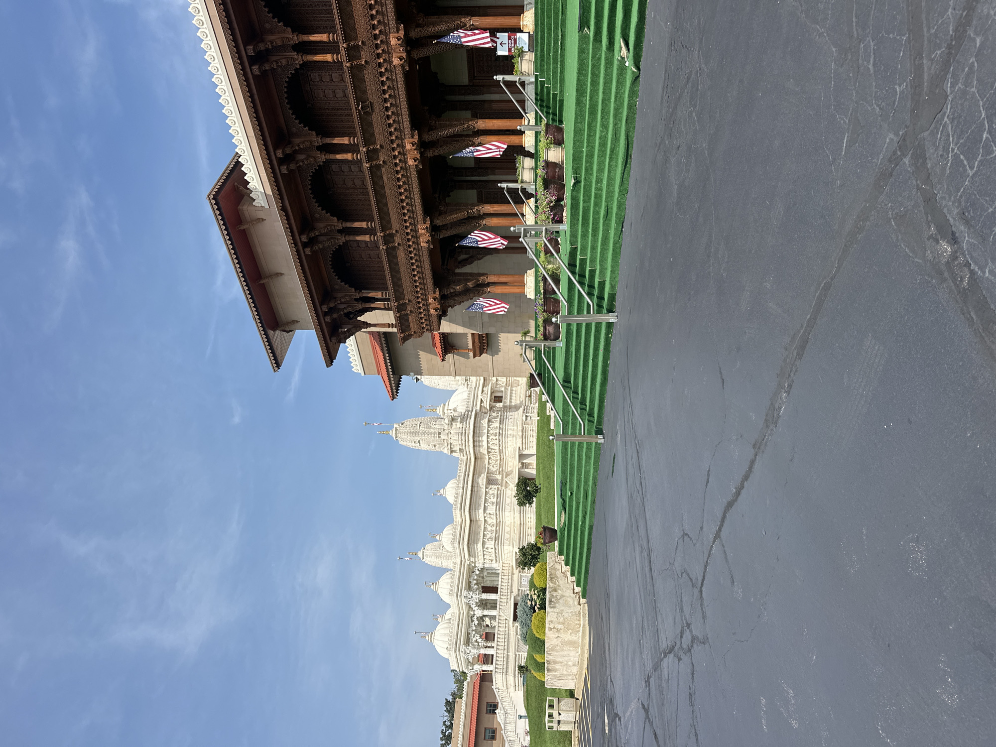 The image shows a view of the BAPS Shri Swaminarayan Mandir in Bartlett, Illinois, USA. The foreground features a cracked asphalt parking lot leading to a set of green-carpeted stairs with metal railings. American flags are displayed on poles along the stairs. To the right of the stairs, a portion of a building with ornate wooden architecture and a red roof is visible. In the background, a large, white marble temple structure with multiple spires and intricate carvings dominates the scene. The sky is blue with scattered white clouds. The environment appears to be outdoors on a clear day.