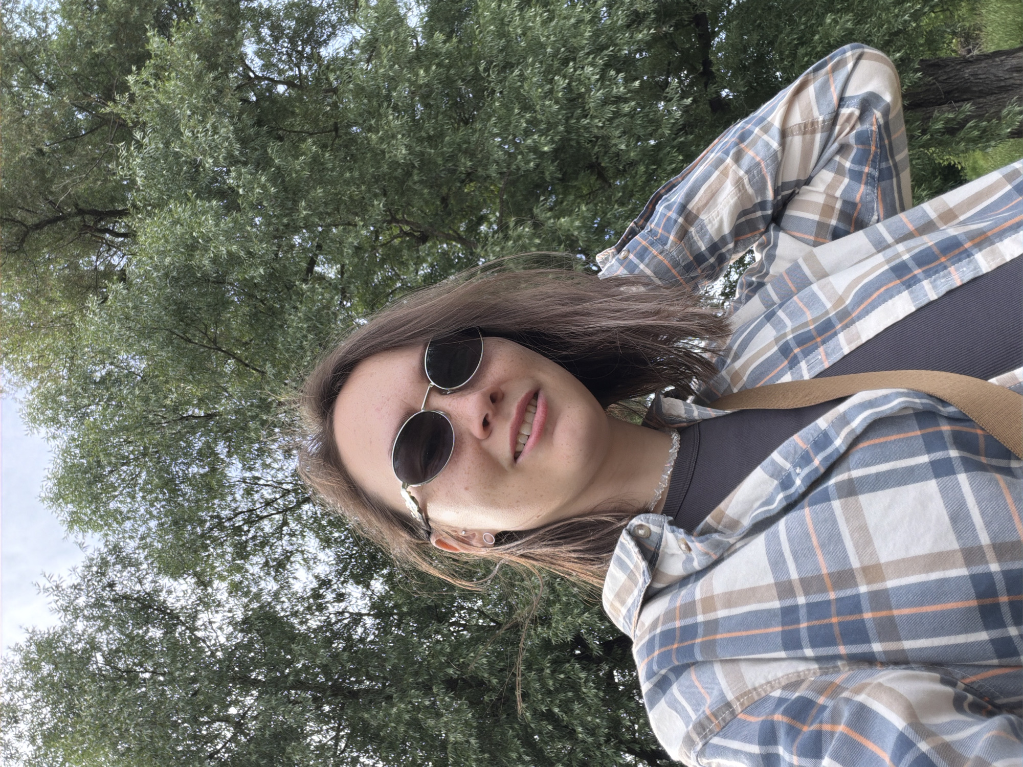 In Garevaya, Russia, a young woman with medium-length brown hair, wearing sunglasses and a plaid shirt, stands outdoors. She is smiling and touching her hair with her right hand. A strap from a bag is visible across her chest. The background is dominated by a large tree with green leaves and a cloudy sky.