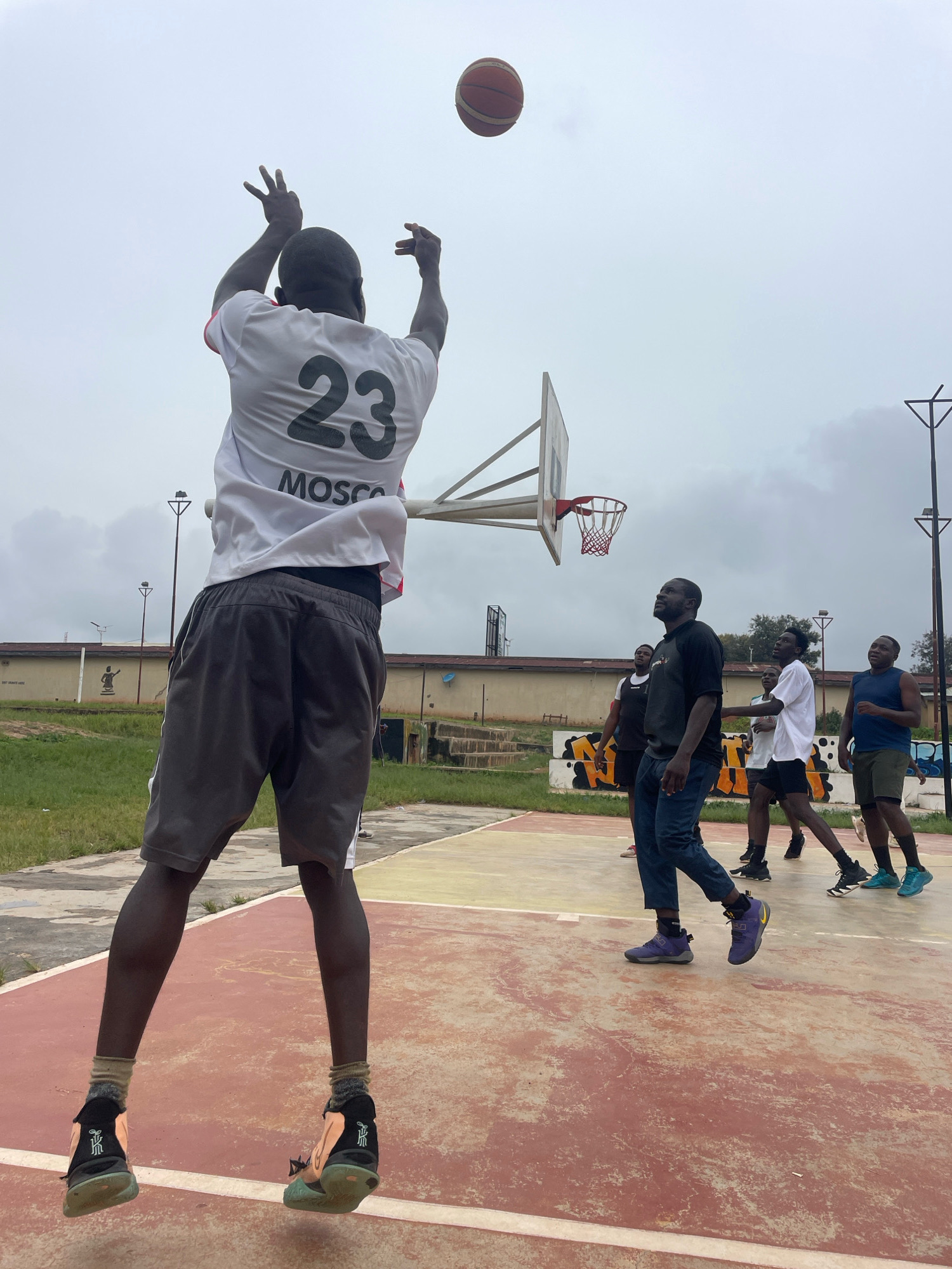 In Gura Top, Nigeria, on an overcast day, several men are playing basketball on an outdoor court. The court surface is a faded reddish-pink and yellow. A basketball hoop with a white backboard and red net stands prominently. In the foreground, a man wearing a white t-shirt with the number "23" and "MOSCO" printed on it, along with grey shorts, is in mid-air, shooting the basketball. He is wearing black and orange basketball shoes. Other men are scattered on the court, some watching the shot, others in defensive or offensive positions. They are dressed in casual athletic wear. Behind the court, a low-rise building with a beige facade and some graffiti is visible, along with grassy areas and trees. Lampposts are also present on the perimeter of the court.