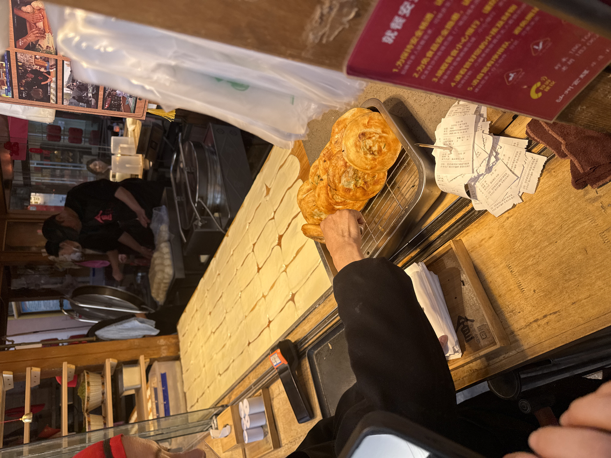 A photo captures an inside view of a food stall in Dunhuang City, China. A person wearing black is reaching into a metal rack full of golden brown, round pastries. To the left, a long counter is filled with sheets of rolled dough. In the background, other people are visible behind a glass-enclosed area. Various receipts and a brown towel sit on the counter to the right. Above, in the top right corner, a red sign with Chinese text is displayed. There are photos hanging on the left wall.