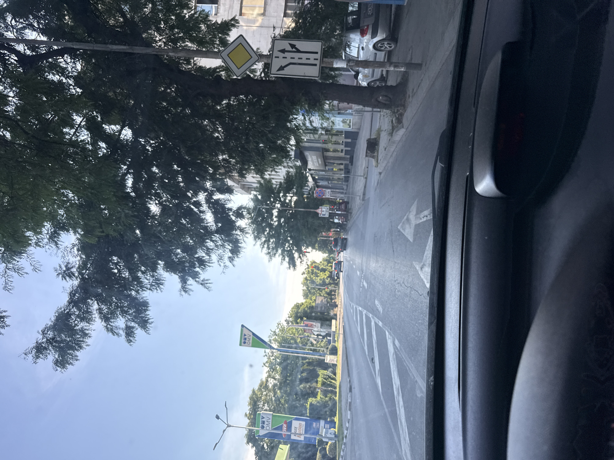 The image shows a street view from a car in Stara Zagora, Bulgaria, on a sunny day. The road has multiple lanes with white markings. On the left side of the road, there is a gas station with OMV branding. Further down the street, there are buildings and trees lining the sidewalk. Traffic signs are visible, including a diamond-shaped yellow sign and a sign indicating lane directions. Cars are present on the road, and a silver car is parked on the right side. The sky is clear blue with some white clouds. The foreground shows the dashboard of the car from which the image is taken.