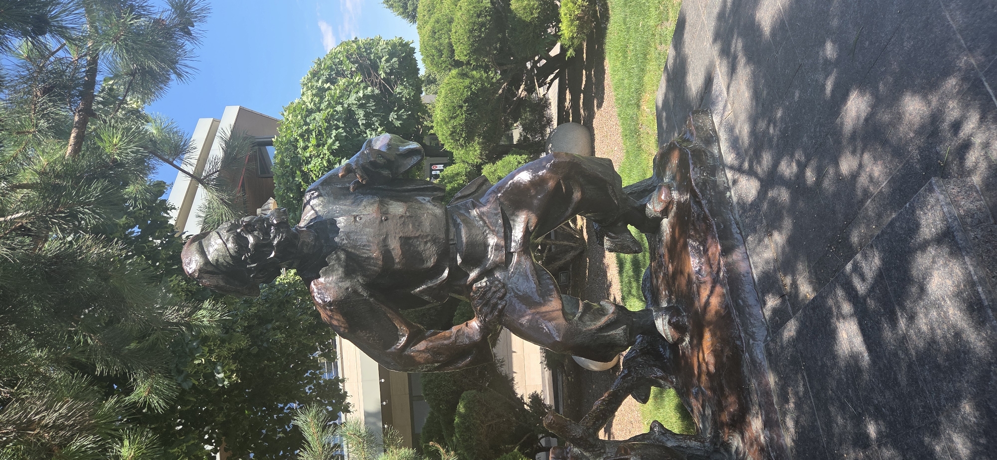 The image shows a bronze statue of a man in an outdoor setting, possibly a park or public square. The statue depicts a man with a beard, wearing a hat, vest, and pants, with his hands on his hips and one leg extended. The statue is dark brown and appears to be weathered, with a shiny patina in some areas. It is placed on a rough, dark-colored base that resembles rock or earth.
In the background, there are lush green trees and bushes, as well as a modern building with a light-colored facade and large windows. The sky is blue with a few white clouds, suggesting a clear, sunny day. The ground in front of the statue is paved with dark, textured tiles, and dappled shadows from the trees are cast across the surface.
This statue is located in Rostov-on-Don, Russia, specifically in the park area adjacent to the Don State Technical University. The statue is of a Cossack.
