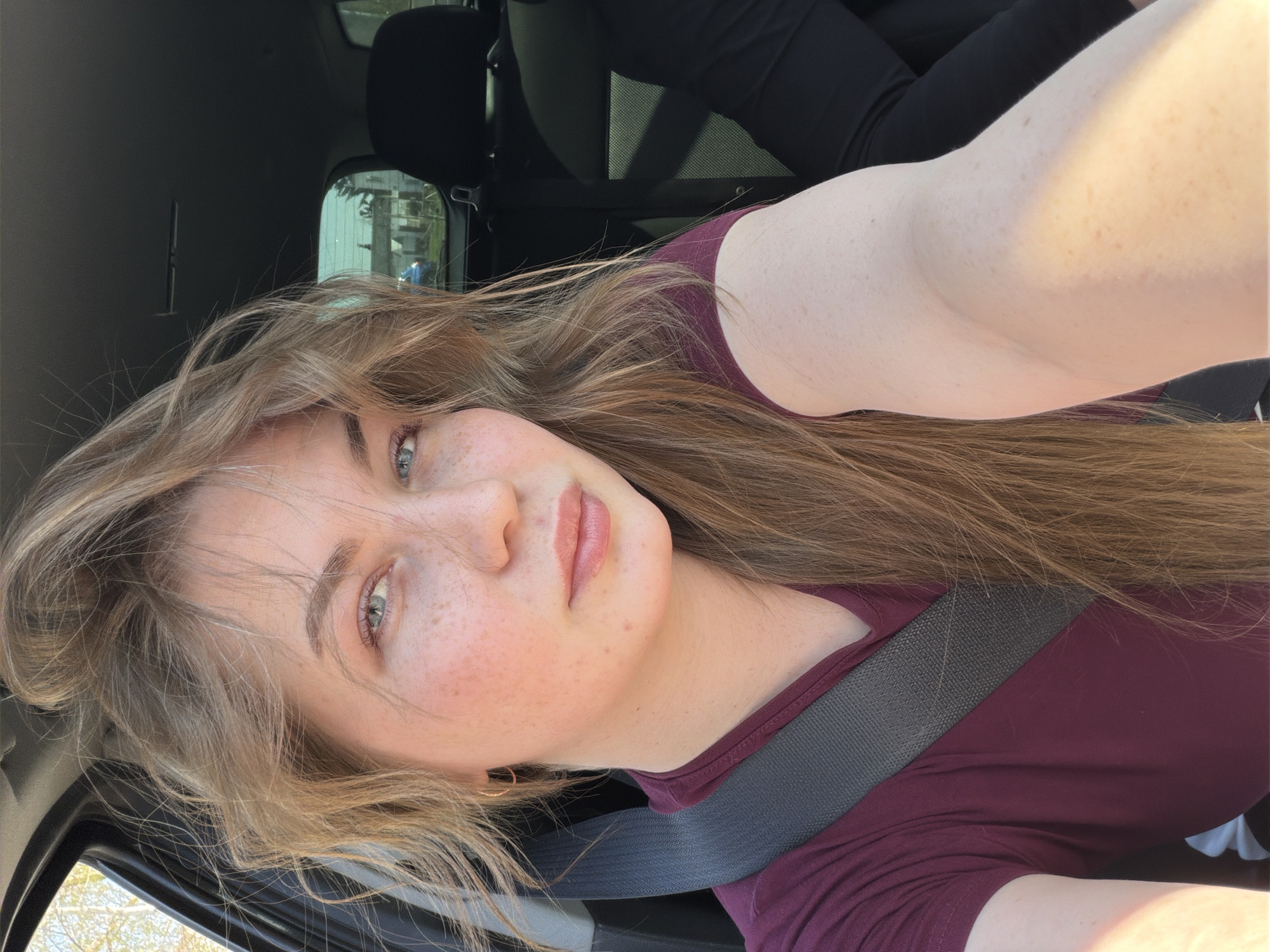A light-skinned young woman with light brown hair and freckles is wearing a maroon shirt and a seatbelt in a car. The car's interior is dark. Outside the window, blurry buildings and trees are visible, but the specific location in the city of Chaikovsky, Russia, cannot be determined from the visual information.