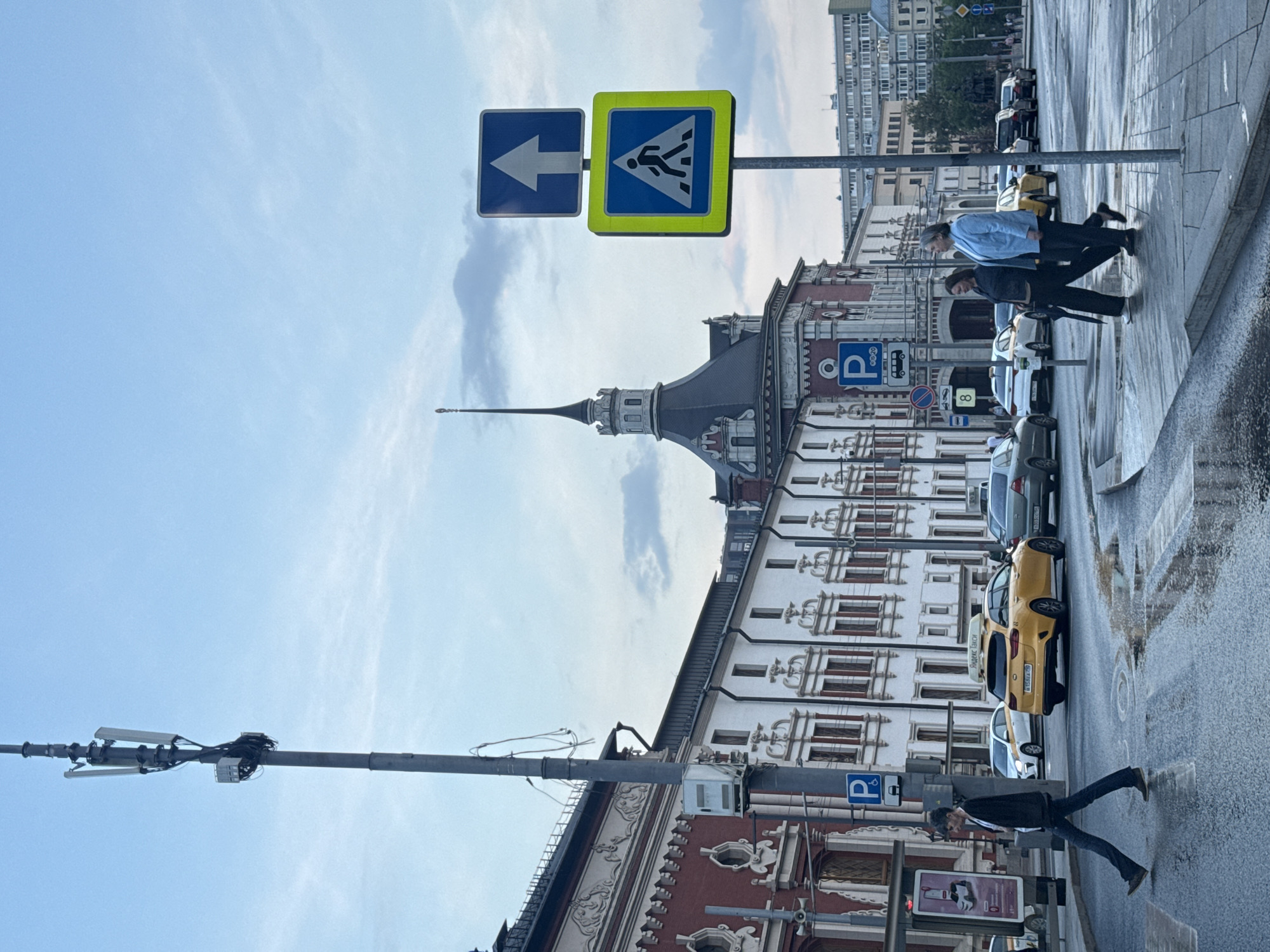The image shows a street scene in an urban environment. In the foreground, a crosswalk sign with an arrow pointing left and a pedestrian crossing sign are visible. Two individuals are walking on the sidewalk, while parked cars and a yellow taxi line the street. The architecture in the background features a distinctive building with a pointed spire and detailed facade. A utility pole with mounted equipment is visible extending into the sky. The street appears wet. The location of Ukromne, Ukraine, is not verifiable based on image data.