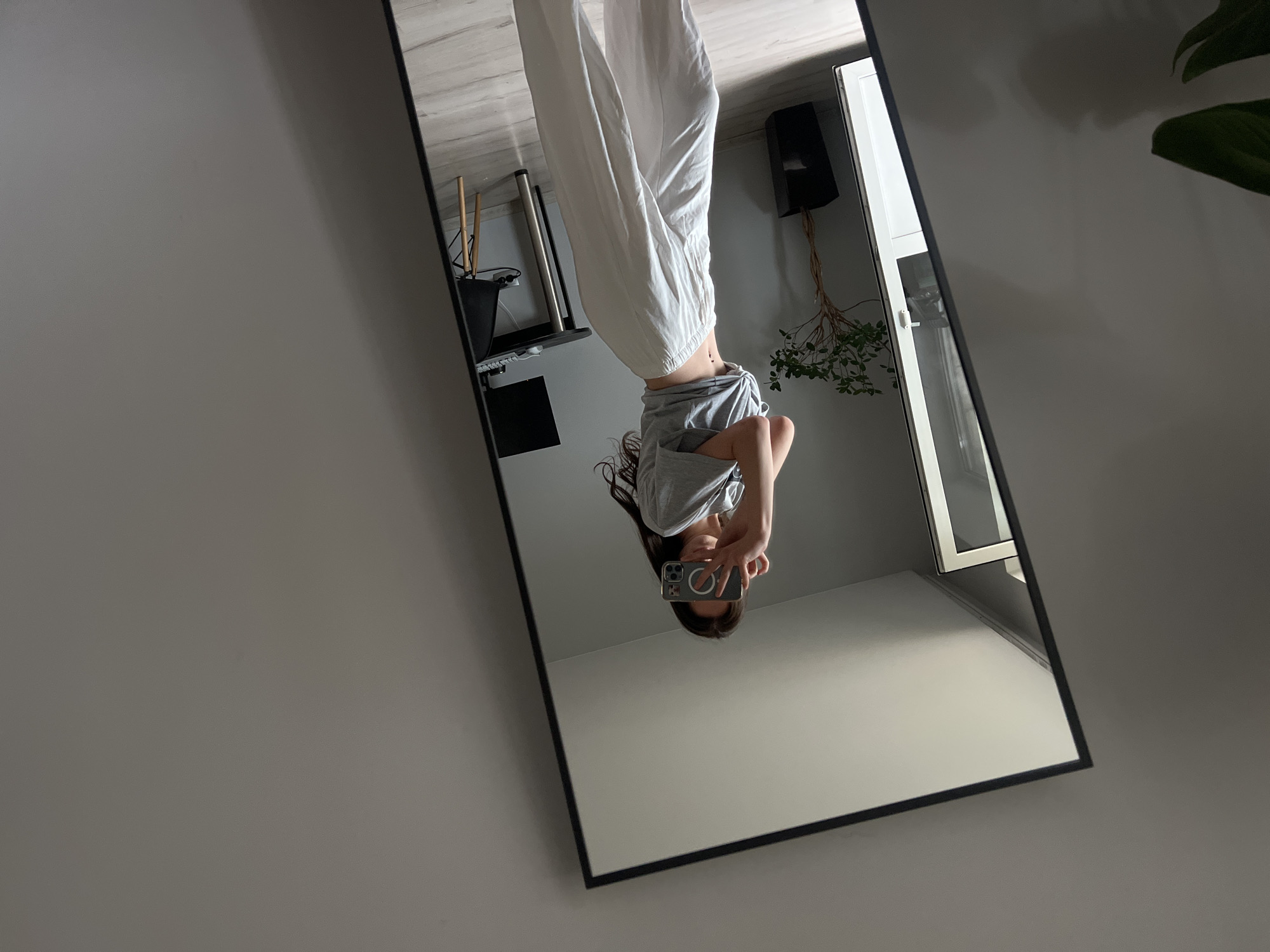 A full-length mirror with a black frame captures a reflection of a young woman taking a mirror selfie. The original image file is rotated counter-clockwise, making the reflection appear upside down. In the reflection, the woman is seen holding a smartphone with her right hand, obscuring her face. She wears a plain grey t-shirt and loose-fitting white pants, with her dark hair falling around her shoulders. The back of the smartphone has a distinctive white 'O' shaped design on a black accessory.
The background reflected in the mirror reveals an indoor room with light-colored walls and ceiling. To the left side of the reflection, a black bin or pot holds several light-colored wooden sticks. Further into the reflection, a black, possibly wall-mounted, speaker is visible above a plant with green leaves and exposed roots trailing downwards. A white-framed window or door is also reflected on the right. The portion of the wall outside the mirror, which borders the left side of the frame, is a plain light gray. The scene is illuminated by soft, natural light, indicating it is daytime. The location is Ternopil, Ukraine.