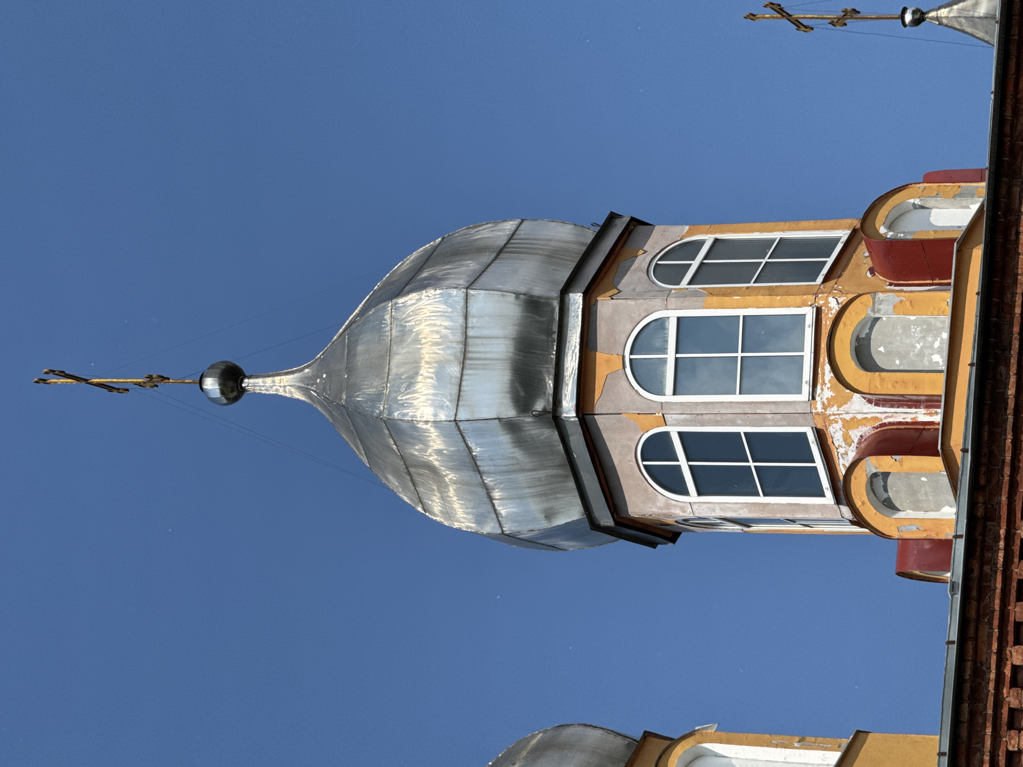 This is a close-up, low-angle, outdoor, daytime shot of the onion dome and upper facade of a religious building in Chudovo, Russia. The sky is a clear, bright blue. The main focus is on a large, silver-colored onion dome with a golden cross at its apex. The dome is constructed of segmented metal panels and shows signs of weathering and wear. Below the dome is a section of the building's facade painted in a faded yellow and orange color, with patches of peeling paint revealing a lighter colored underlayer. There are arched windows with white frames on this level, two of which are clearly visible. To the left, a portion of another, smaller silver onion dome is visible. The lower part of the image shows a dark red brick structure with a metallic roof. The overall impression is of an aging but still majestic religious structure against a clear sky. There are no people or animals visible in the frame.