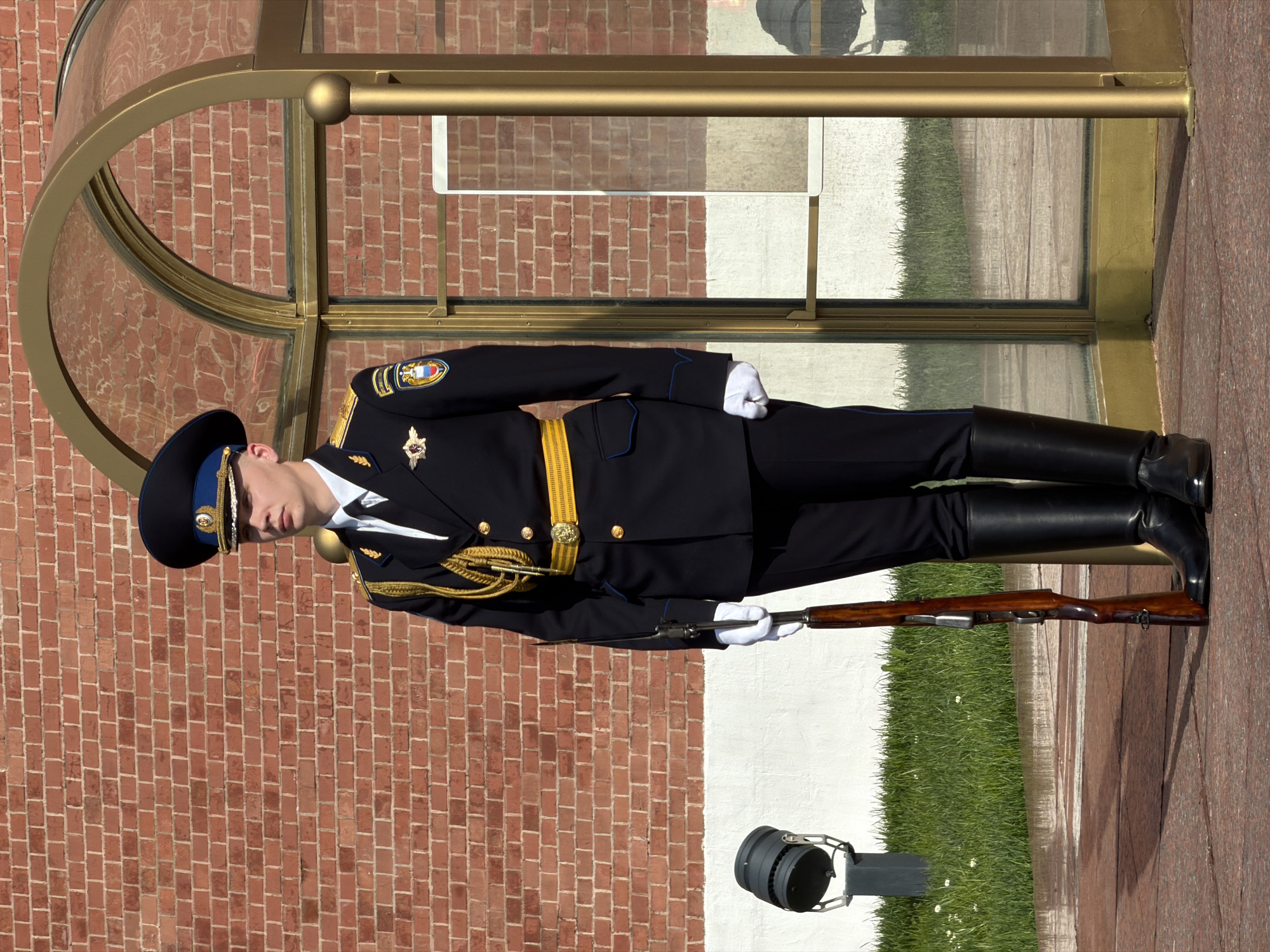 A photograph depicts a Russian honor guard standing at attention in front of what appears to be a glass-enclosed entryway constructed with brick walls. The guard is wearing a black uniform with gold embellishments, including a peaked cap with gold trim. They are holding a rifle, and wearing white gloves. The surrounding environment features a combination of brick and green foliage.