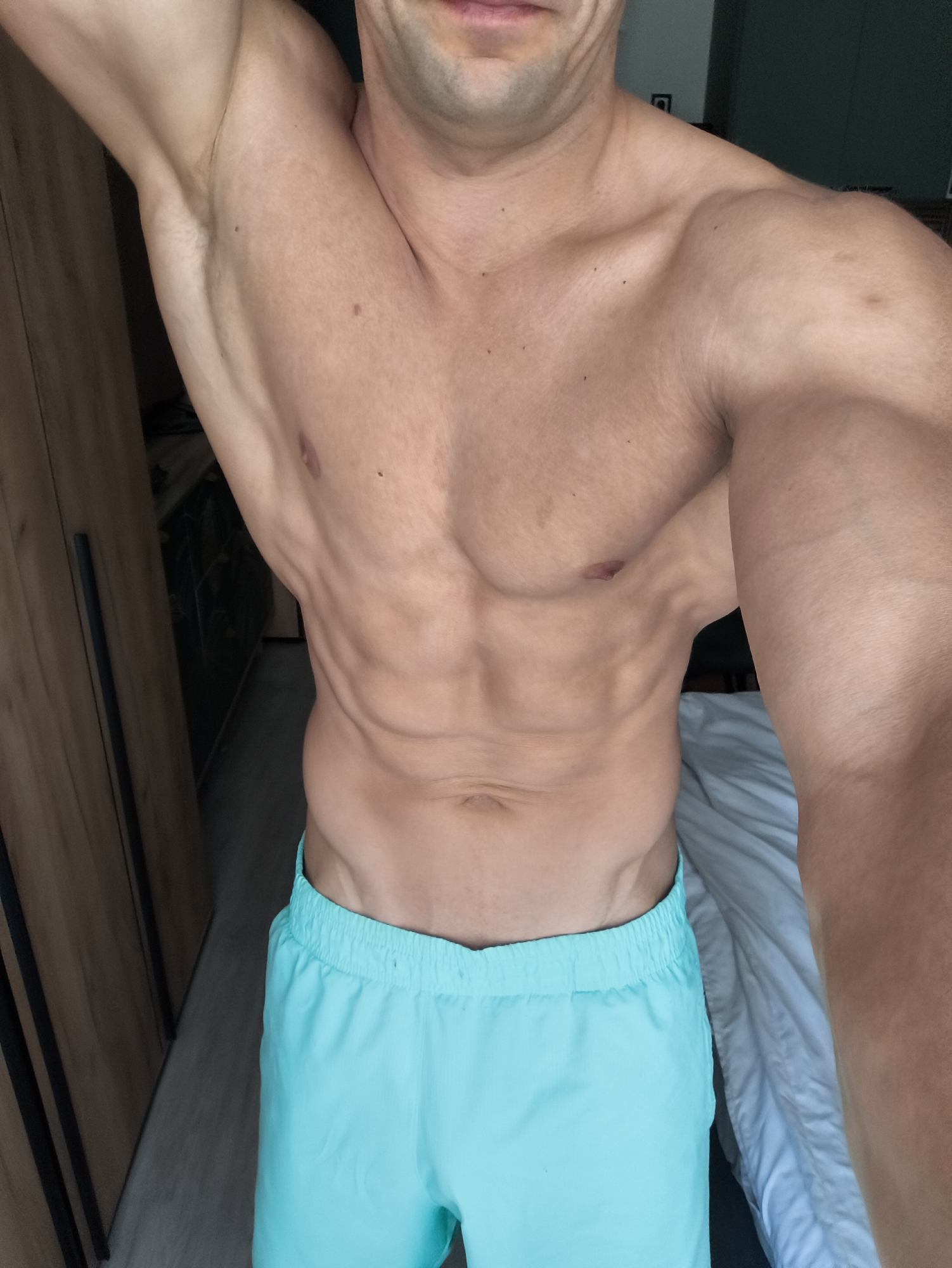 A man is standing in a room. He is shirtless and wearing blue shorts. He has defined abdominal muscles. The room appears to be a bedroom, with a bed visible in the background and a wardrobe on the left. The exact location within Moscow, Russia is indeterminable from the image.