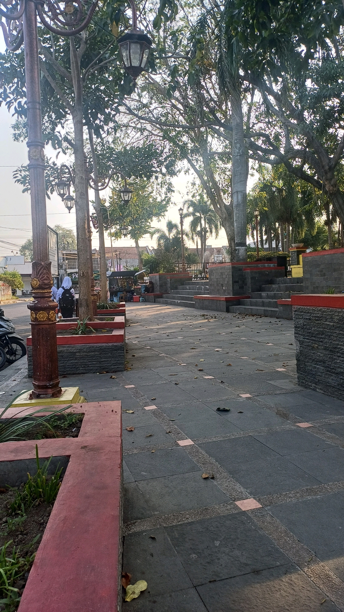 A paved walkway in a park-like setting is depicted. The walkway is made of dark gray stone tiles with decorative pink square inserts. The path is bordered by a red-painted concrete planter box on the left, which contains some green foliage. Further along the walkway, there are tiered stone steps with red accents, suggesting a gathering or seating area.
Several trees with dense foliage are present, casting shadows on the walkway. Decorative lampposts with ornate designs and dark lanterns are spaced along the path. In the background, there are buildings and a street with a parked motorcycle.
On the left side of the walkway, near a lamppost, a person wearing a white hijab and a backpack stands facing away from the camera, partially obscured by a tree. A motorcycle is parked on the street to the left of the walkway. Further down the walkway, there are small market stalls and a few people seated. The overall atmosphere appears calm, possibly in the late afternoon due to the lighting.
While the image is set in a park-like environment, specific details linking it to Klaten, Indonesia, are not directly visible. However, the architectural style of the lampposts and the general streetscape are consistent with urban environments in Indonesia.