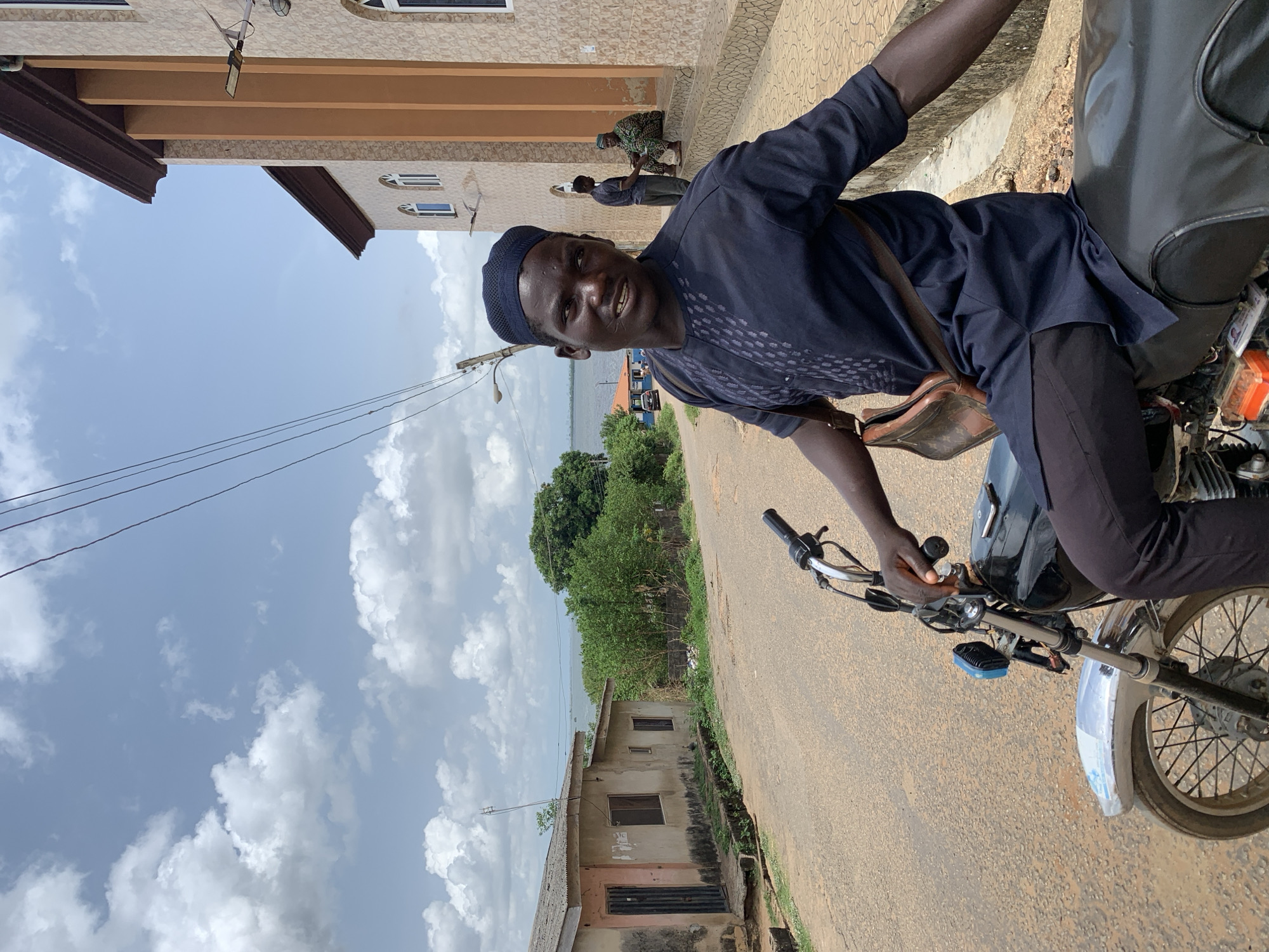 A man rides a motorcycle down a street in Ikorodu, Nigeria. He wears a dark blue patterned shirt and pants with a matching kufi. He has a tan leather bag slung over his shoulder. The street is paved and bordered by buildings, including a two-story beige structure with brown trim and a dilapidated one-story building. A group of people are standing near the two-story building. A body of water is visible in the distance beyond some trees. The sky is light blue with scattered clouds. Overhead power lines stretch across the sky.