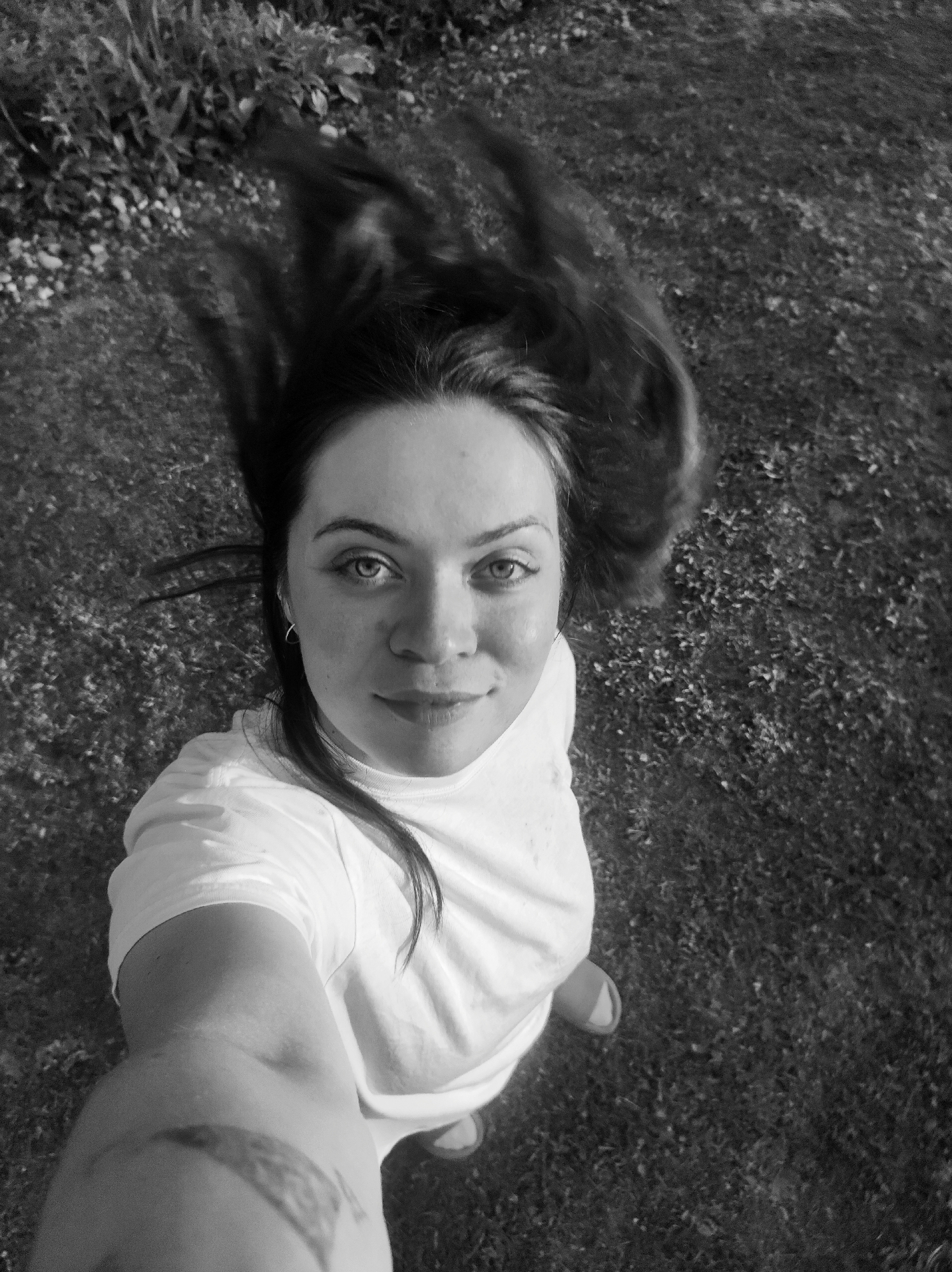 A monochromatic, high-angle selfie depicts a woman with dark hair and light eyes looking directly at the camera. Her dark hair appears in motion, flowing upwards and outwards from her head. She wears a plain, light-colored (likely white) short-sleeved t-shirt. A small hoop earring is visible in her right ear, and a tattoo or distinct marking is present on her right forearm/upper arm. The background consists of a grassy area with some sparse, low-lying vegetation, and denser foliage towards the upper left. The scene is outdoors under natural daylight, located in the city of Фоки, Russia.
