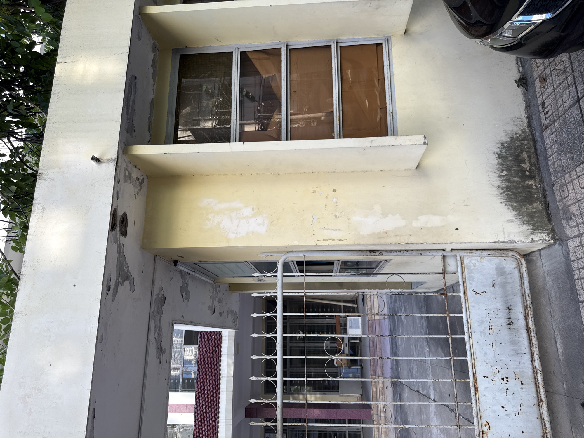 The image shows a weathered, off-white metal gate in front of a building with a yellow facade. The gate is partially open, revealing a courtyard with a paved surface and some indistinct objects, possibly furniture. Above the gate, there is an opening into the building, showing a glimpse of a tiled roof and a tall, multi-story structure with windows. To the right of the gate, a large window with brown-tinted glass is visible on the yellow wall. The building appears to be in a state of disrepair, with peeling paint and water stains on the facade. Green foliage is visible at the top of the image, suggesting the presence of trees or plants. A portion of a black car is visible in the lower right corner. The scene is set in a city, likely in Nha Trang, Vietnam, given the architectural style and the overall appearance of the building and its surroundings. The environment appears to be a street or a public area adjacent to the building.