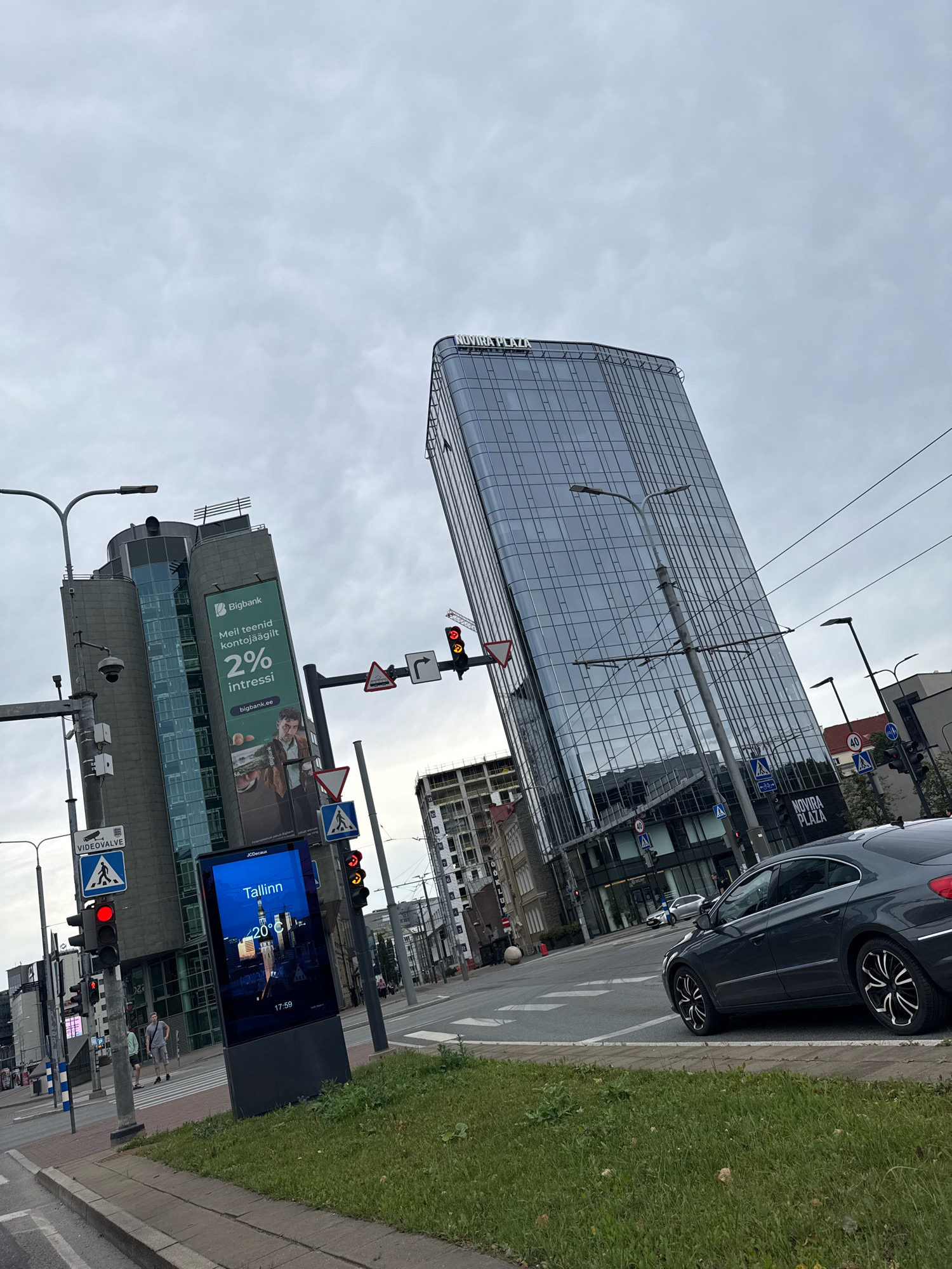 A wide-angle, slightly tilted shot captures an overcast street scene in Tallinn, Estonia, dominated by modern architecture. In the foreground, a dark gray sedan is parked on the right side of the road, its wheels turned slightly. To the left, a gray, cylindrical building with a glass facade displays a large green billboard advertising "Bigbank" with an offer of "2% interest." Below this, a digital screen shows a nighttime view of Tallinn with the temperature at 20°C and the time 17:59. A traffic light for vehicles is red, with pedestrian crossing signals also visible. Further back, a tall, sleek glass skyscraper with "NOVIRRA PLAZA" visible at its base stands prominently. Several other buildings, construction sites, streetlights, and traffic signs are scattered throughout the urban landscape. Two men are visible walking on the sidewalk near the pedestrian crossing. The sky is filled with gray clouds.