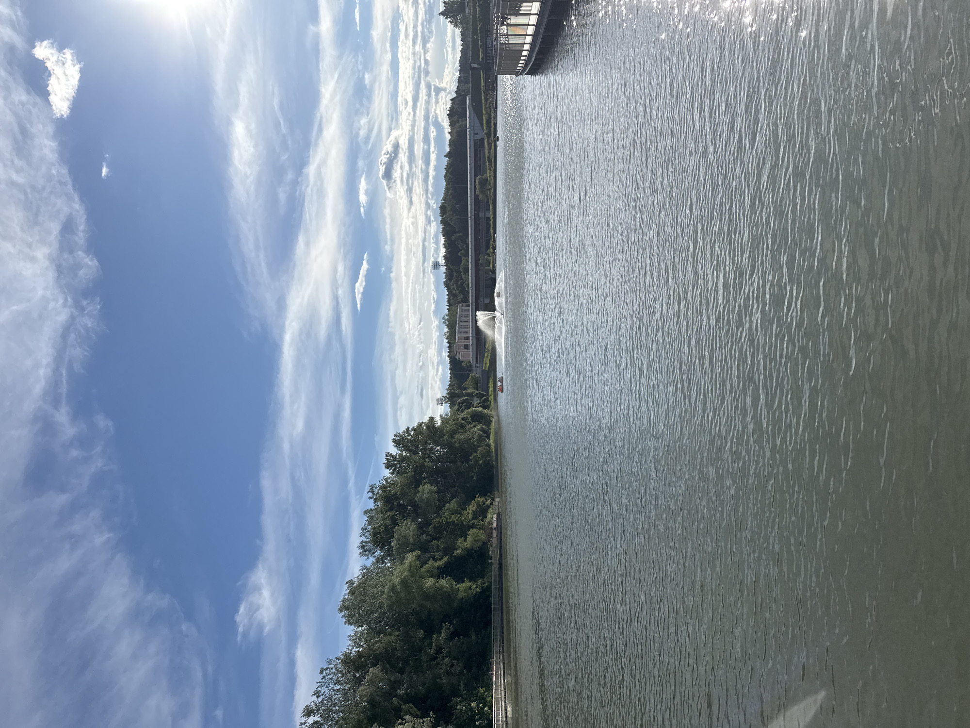 The image shows a serene lake in Zelenograd, Russia. On the left side of the frame, a dense green forest borders the lake, with a wooden pier extending partially into the water. In the distance, a bridge spans across the lake, and a building with a fountain is visible. On the right side of the image, another wooden pier with railings is present, suggesting a recreational area. The sky is a vibrant blue, dotted with white clouds, and the sun is shining brightly, casting reflections on the water's surface. A small boat with two occupants can be seen on the lake, enjoying the scenery. The overall scene depicts a peaceful day at a park or resort area.