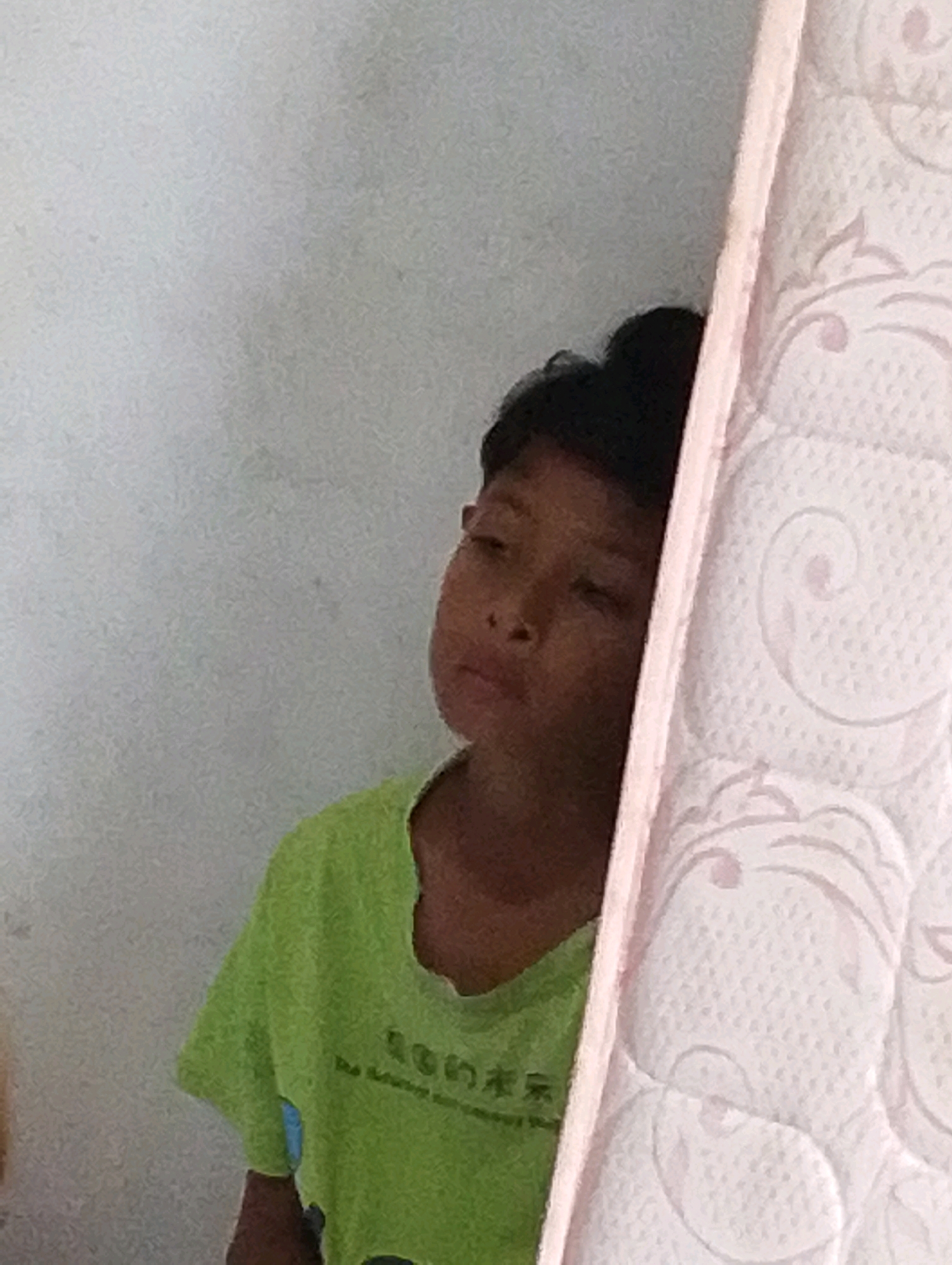 A young child, appearing to be of Southeast Asian descent, is partially visible in the frame, positioned to the left of a light pink, patterned object, possibly a mattress. The child is wearing a faded lime green t-shirt with some Asian characters printed on it. Their head is tilted slightly downwards, and their eyes are looking towards the left. The background is a plain, textured light grey wall. The scene appears to be indoors, in Manaruwi, Indonesia.