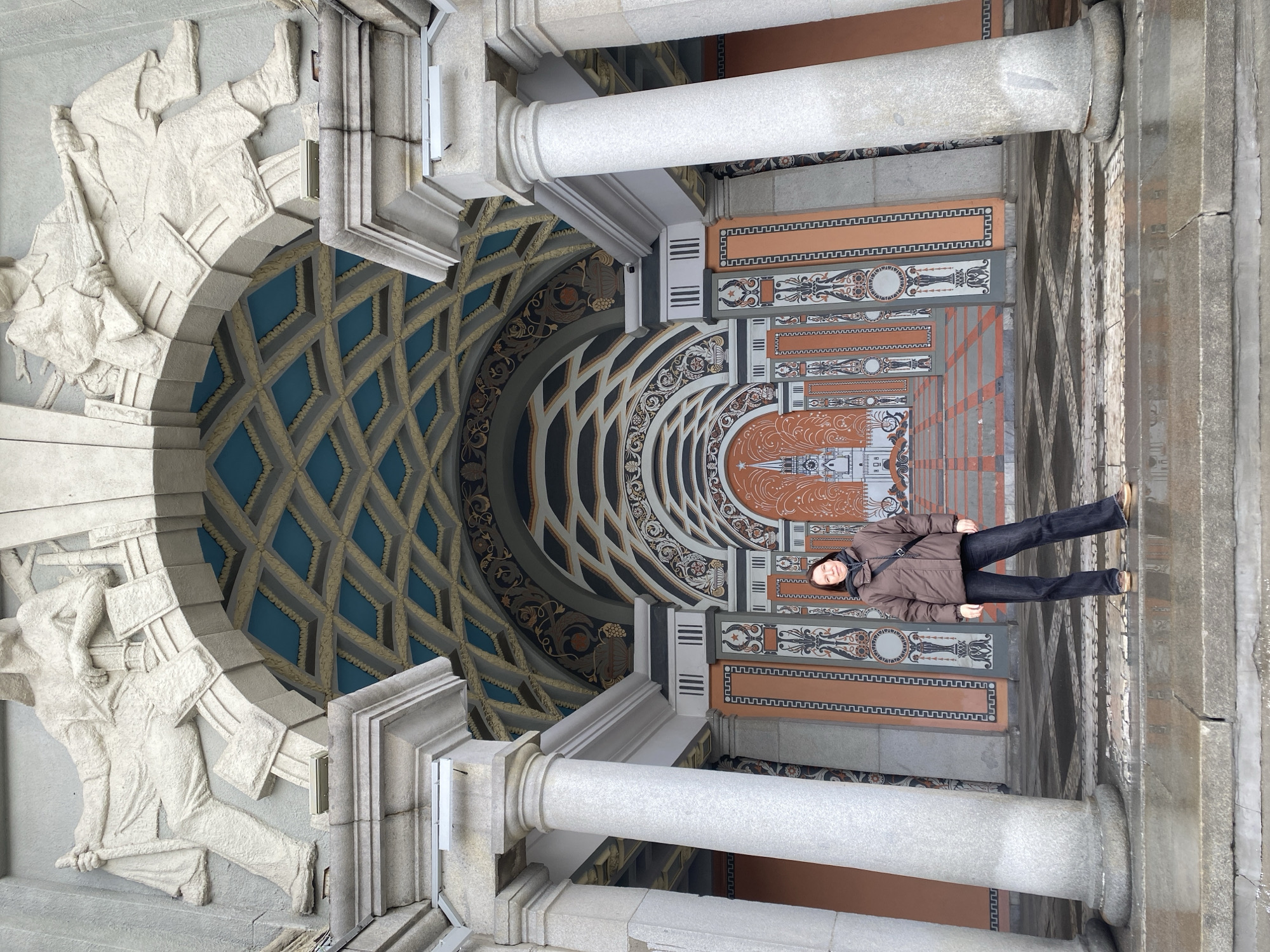 A woman is standing in a corridor or entrance area of a building in Yekaterinburg, Russia, as the decorations are representative of the architectural style of the city. The corridor features a decorative ceiling with a geometric pattern in blue and gold, an archway with sculpted figures, and columns. The floor is patterned with dark lines. The woman is wearing a brown jacket and dark pants. She is standing casually with her hands at her sides and looking toward the camera. The lighting appears to be natural daylight. The scene is captured from a slightly low angle.