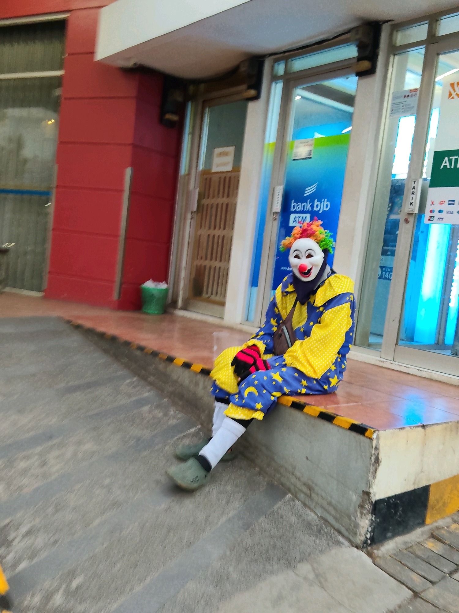 In Purwakarta, Indonesia, a person dressed as a clown sits on a ledge outside an ATM. The clown wears a white mask with a red nose, a colorful wig, and a blue and yellow costume adorned with yellow stars. The costume includes a yellow shirt with blue polka dots on the sleeves and collar, and blue pants with yellow stars. The clown also wears white socks, green shoes, and red gloves. Behind the clown are the glass doors of an ATM, with a blue sign displaying "bank bjb ATM". The environment appears to be a public area, possibly near a building with a red facade. The ground is a mixture of concrete stairs and a paved walkway. A green trash bin is visible to the left of the ATM. The overall scene is captured in a slightly blurry, candid manner.