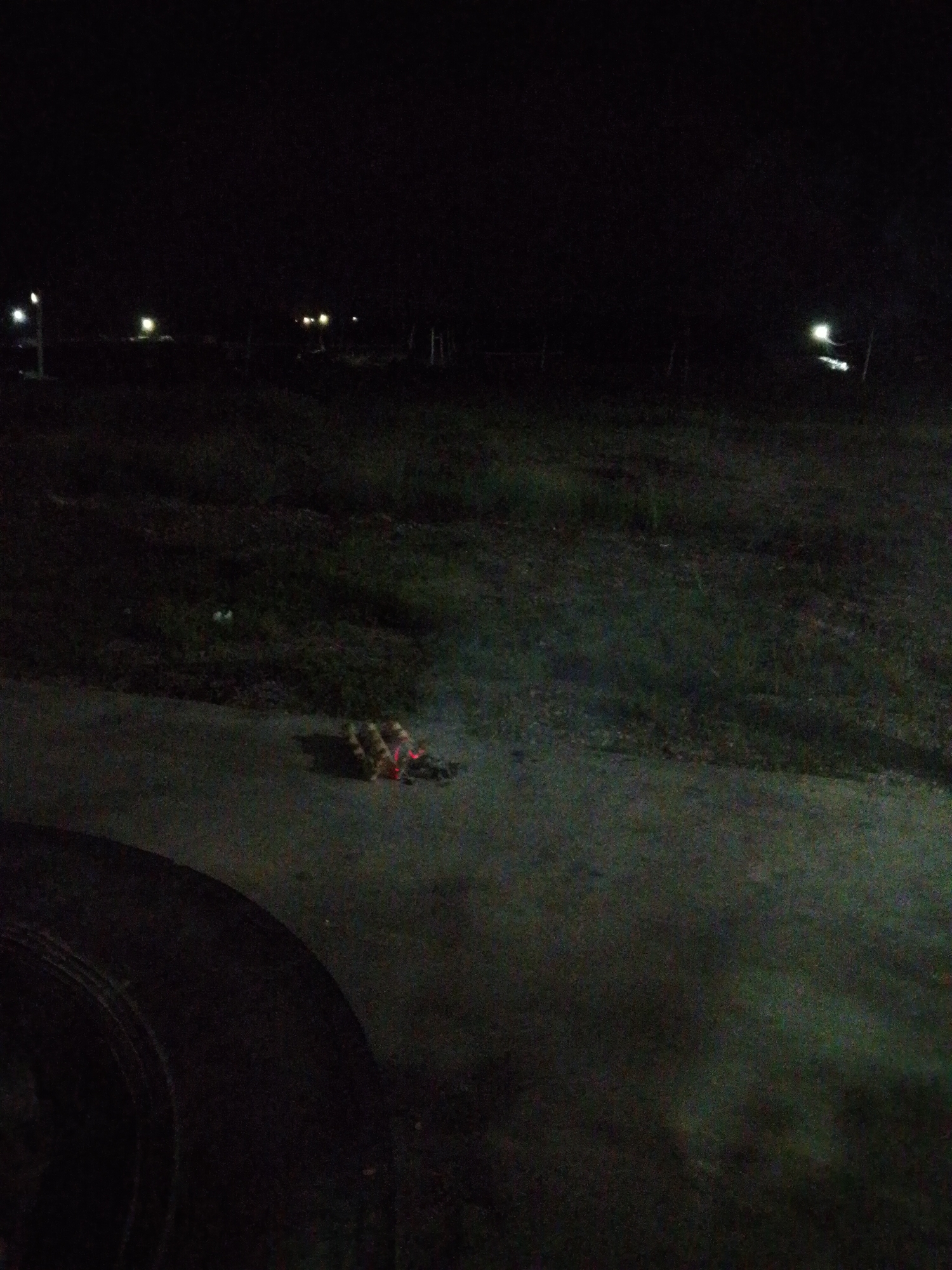 The image shows an outdoor scene at night, likely in a sparsely populated area. There is a low road or paved surface in the foreground. A tire is visible in the lower left corner. In the middle of the paved area, there is what appears to be a cluster of burning or smoldering debris. In the background, there is a dark, grassy area with scattered lights. The environment suggests an area in or near Bukit Ketok, Indonesia, with some artificial illumination in the distance. The specific details of the burning debris are unclear due to the darkness and image quality.