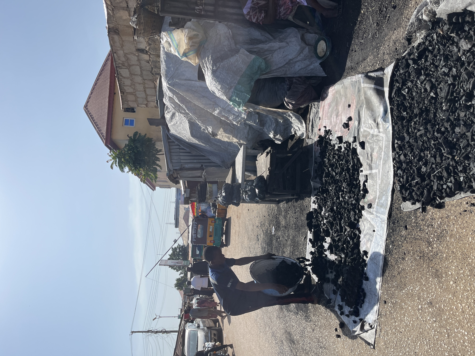 The image depicts an outdoor scene in the city of Gura Top, Nigeria, likely a market or roadside stall. A young man, wearing a dark blue t-shirt with a logo and red trim on his pants, is actively pouring charcoal from a large metal basin onto a white tarp spread on the asphalt road. The charcoal is dark and fragmented, scattered across the tarp and the ground.
In the background, several other individuals are visible. To the left, people are walking along the road, with cars and a three-wheeled vehicle also present, suggesting a busy thoroughfare. To the right, a stall is set up with a large tarp covering goods, possibly charcoal or other market items. A few people are seated near the stall, one woman wearing a patterned headwrap and dress. The environment appears to be sunny, with a clear blue sky. Buildings and power lines are visible in the distance. The overall impression is of a bustling commercial area, with a focus on the preparation or sale of charcoal.