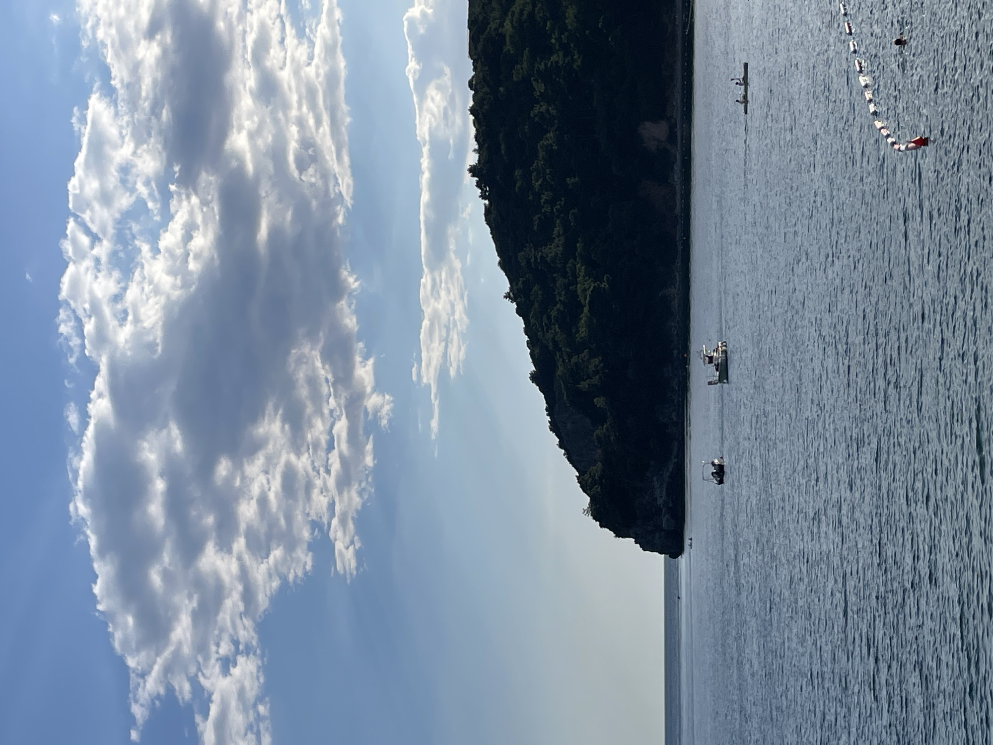 The image shows a view of the sea in Budva, Montenegro. A dark, green, tree-covered cliff is at the edge of the water. The sky is a clear, light blue with fluffy, white clouds, and there are a few boats on the water. There is a canoe or kayak to the right of the frame in the water as well as a barrier with alternating red and white rectangular floats.