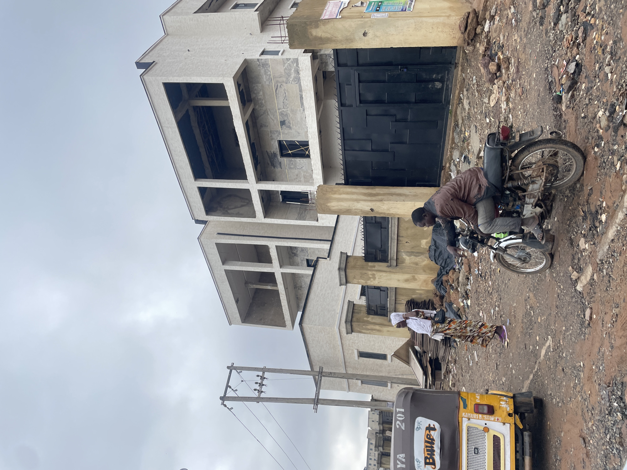 This is a ground-level, medium shot of a construction site in Guzape, Nigeria. In the foreground, a yellow and brown auto-rickshaw, branded "Bullet," is parked on the left side of the frame. To the right of the auto-rickshaw, a woman wearing a white headscarf and a patterned skirt stands looking to her right. A man is seated on a motorcycle to the right of the woman, facing away from the camera. The background features a multi-story building under construction, with unfinished facades and open window frames. A large black metal gate is visible in front of the building. The ground is dirt and gravel, with scattered debris, including planks of wood and rubble. The sky is overcast and a utility pole with wires stands to the left of the building.