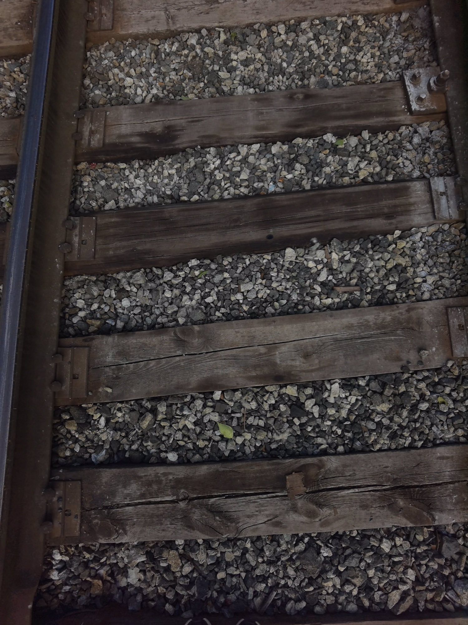 The image shows a close-up, overhead view of a section of railroad tracks. The tracks consist of a steel rail on the left side and wooden railroad ties laid horizontally across a bed of gravel. The wooden ties are weathered and appear to be old, with visible grain and some cracks. Metal plates and fasteners are used to secure the rail to the ties. The gravel bed is composed of various sizes of grey and brown stones. The overall scene is one of a functional but aged railroad infrastructure, suggesting it is located in an outdoor, industrial, or transportation-related setting. There are no people or animals visible in the image. Without additional information, it is not possible to determine the specific location in the city of Chaikovsky, Russia.