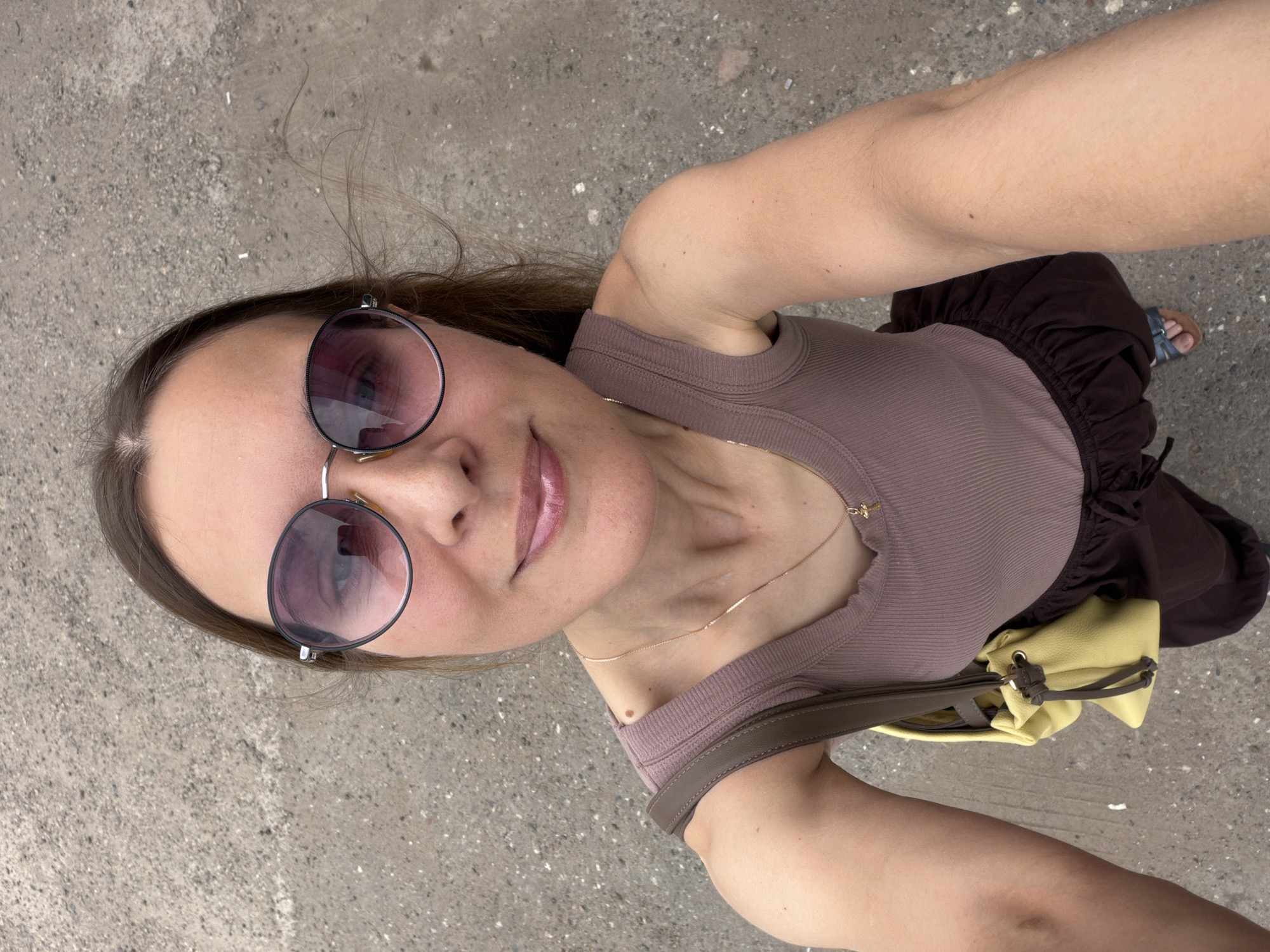 A medium-shot, high-angle selfie shows a woman standing on a concrete surface in the city of Yagul, Russia. The woman has brown hair, is wearing large, round sunglasses with purple lenses, and a mauve ribbed tank top. A delicate gold necklace with a small cross pendant is visible around her neck. A brown strap from a purse is draped over her left shoulder, and a yellow purse hangs in front of her. She is wearing a dark brown tiered skirt and blue and brown sandals. Her left arm is extended, holding the camera. The ground is a rough, gray concrete surface with visible pebbles and small white specks.